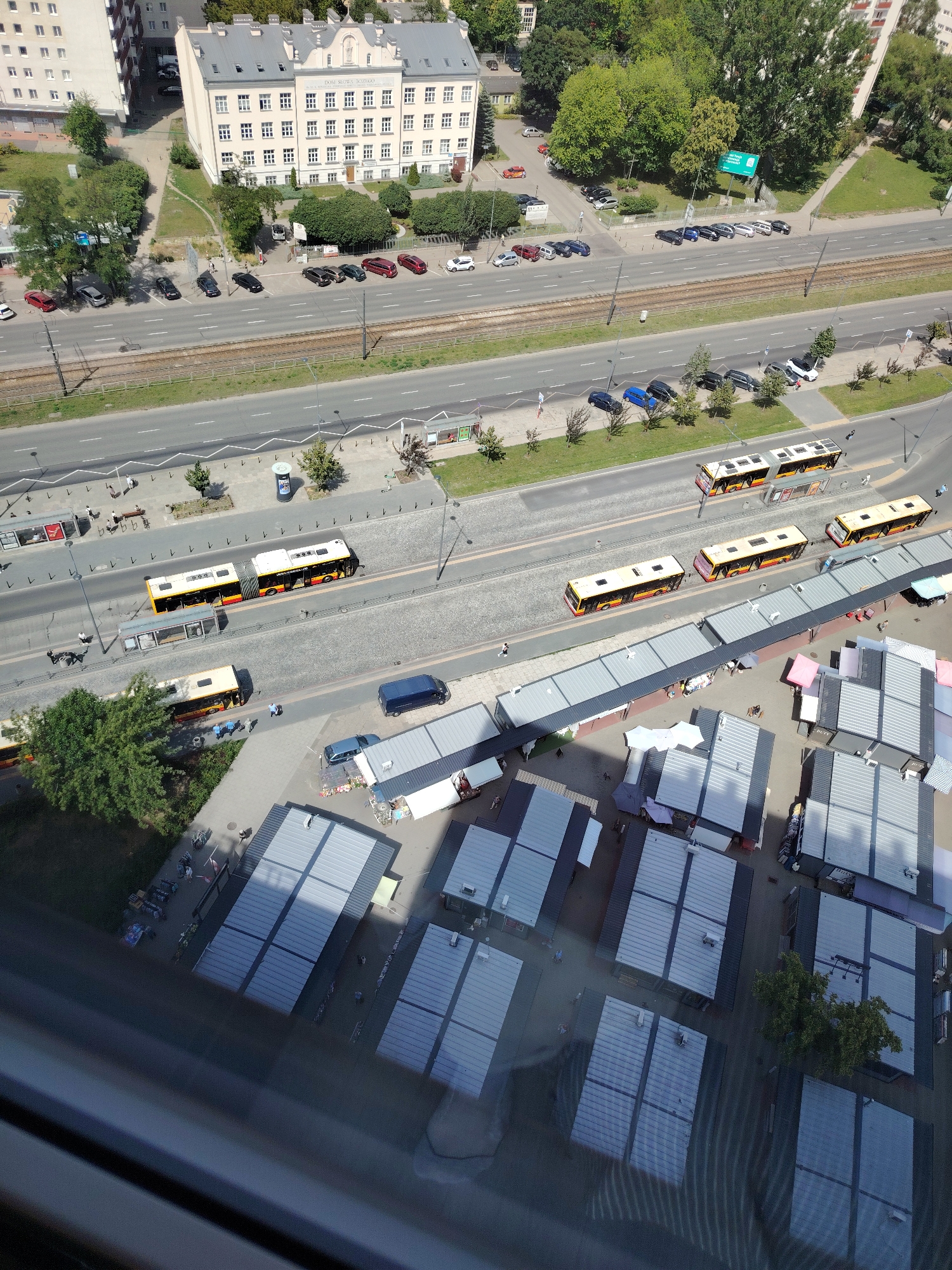 This aerial view captures a busy urban scene in Warsaw, Poland. The image is dominated by a street with several lanes of traffic and a tram track running alongside. Multiple yellow and red buses are stopped at what appear to be bus stops, suggesting a public transportation hub. In the foreground, a market with numerous stalls covered by gray roofs is visible, with a few people browsing or working.
The scene is bathed in bright sunlight, indicating it is daytime, likely midday, given the strong shadows. The buildings in the background are a mix of older, light-colored structures with many windows and a more modern, white building on the left. Green trees are interspersed throughout the urban landscape, adding touches of nature to the built environment.
The activity appears to be normal daily life, with people commuting via public transport and shopping at the market. There are no obvious signs of a specific event taking place, just the general hustle and bustle of a city. The overall atmosphere seems lively and functional.
Notable details include the distinct yellow and red livery of the buses, the organized layout of the market stalls, and the presence of tram tracks, which are common in Warsaw. The reflection on the lower part of the image suggests the photo was taken from a window high up in a building. While some text is visible on the buses and street signs, it is too small to be deciphered. The environment appears to be a public space with commercial and transportation functions integrated.