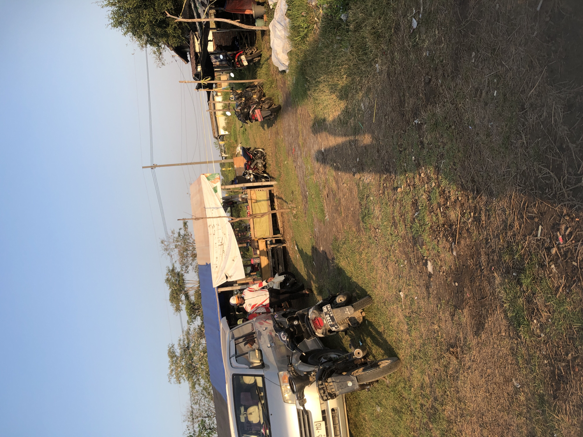This is a forensic, matter-of-fact description of a scene in Gempolsari, Indonesia. The image depicts an outdoor market or roadside stall area during daylight hours, with a clear sky.

In the foreground, the ground is a mix of dry grass, dirt, and debris. Two motorcycles are parked. The one on the left is a dark-colored scooter with its rear wheel prominent. The motorcycle on the right is also dark-colored and has a license plate that reads "W 2280 GK" and "01-25". A silver minivan is parked to the left of the motorcycles. The minivan has a license plate starting with "S 16 - MO".

A man, wearing a white cap, a white jacket with red and black accents, and dark pants, stands near the minivan, facing away from the camera. He appears to be walking or standing by a stall.

Behind the minivan and the man, there is a market stall with a wooden counter and a white canopy supported by poles. Various items are visible on the counter, but they are indistinct. To the right of this stall, there is another structure with a corrugated roof supported by wooden poles. Further to the right, more stalls or structures are visible, including one with a dark roof and a red tarpaulin. Several other motorcycles are parked in front of these structures, some are red and black, others dark.

The environment includes sparse vegetation and trees in the background. There are also utility poles and wires visible against the sky. The shadows of two figures are cast on the ground in the mid-ground, suggesting the presence of at least two people in addition to the visible man. The overall impression is a functional, possibly temporary, commercial area.