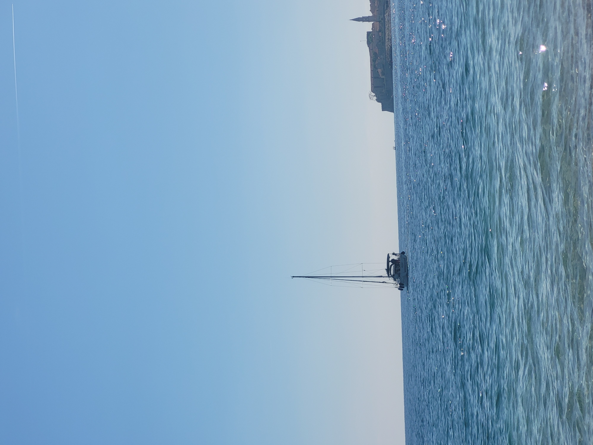 This landscape image depicts a tranquil coastal scene, likely near Budva, Montenegro. The upper two-thirds of the frame show a clear, light blue sky, with a faint white airplane contrail visible in the upper left corner.
The lower third of the image is filled with calm, deep blue water, featuring gentle ripples that reflect sparkling sunlight across its surface. Along the horizon, a distant landmass reveals a historic coastal town, characterized by various buildings including some with distinct spires or bell towers, consistent with European architecture.
A single sailboat is positioned in the water, slightly to the right of the center. It has a tall mast and intricate rigging, with a small cabin or deck area where two small, dark figures appear to be present, possibly seated or standing. No sails are deployed on the boat.
The scene suggests a leisurely activity, with the sailboat possibly anchored or slowly cruising on a peaceful day. The bright sky and glistening water indicate it is daytime under clear weather conditions, evoking a sense of serenity. No visible text is present in the image.