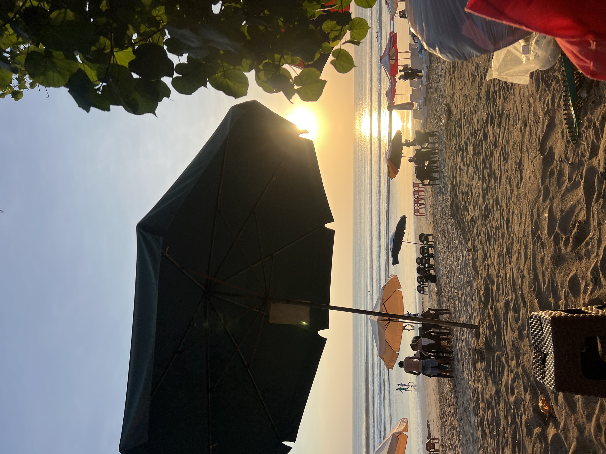 A view of Kuta Beach, Indonesia, during sunset. The foreground is dominated by a large, dark green umbrella, with a smaller, orange and yellow umbrella to its left. Beyond the umbrellas, the sandy beach is populated with several groups of people enjoying the evening. Some individuals are seated at tables under umbrellas, while others are walking or playing near the water's edge. The ocean stretches out to the horizon, with gentle waves rolling in. The setting sun casts a warm, golden glow across the scene, reflecting brightly on the water's surface. In the background, trees with large leaves frame the right side of the image, adding a touch of lush greenery to the coastal landscape. The overall atmosphere is serene and relaxed, capturing the typical evening ambiance of a popular beach destination.