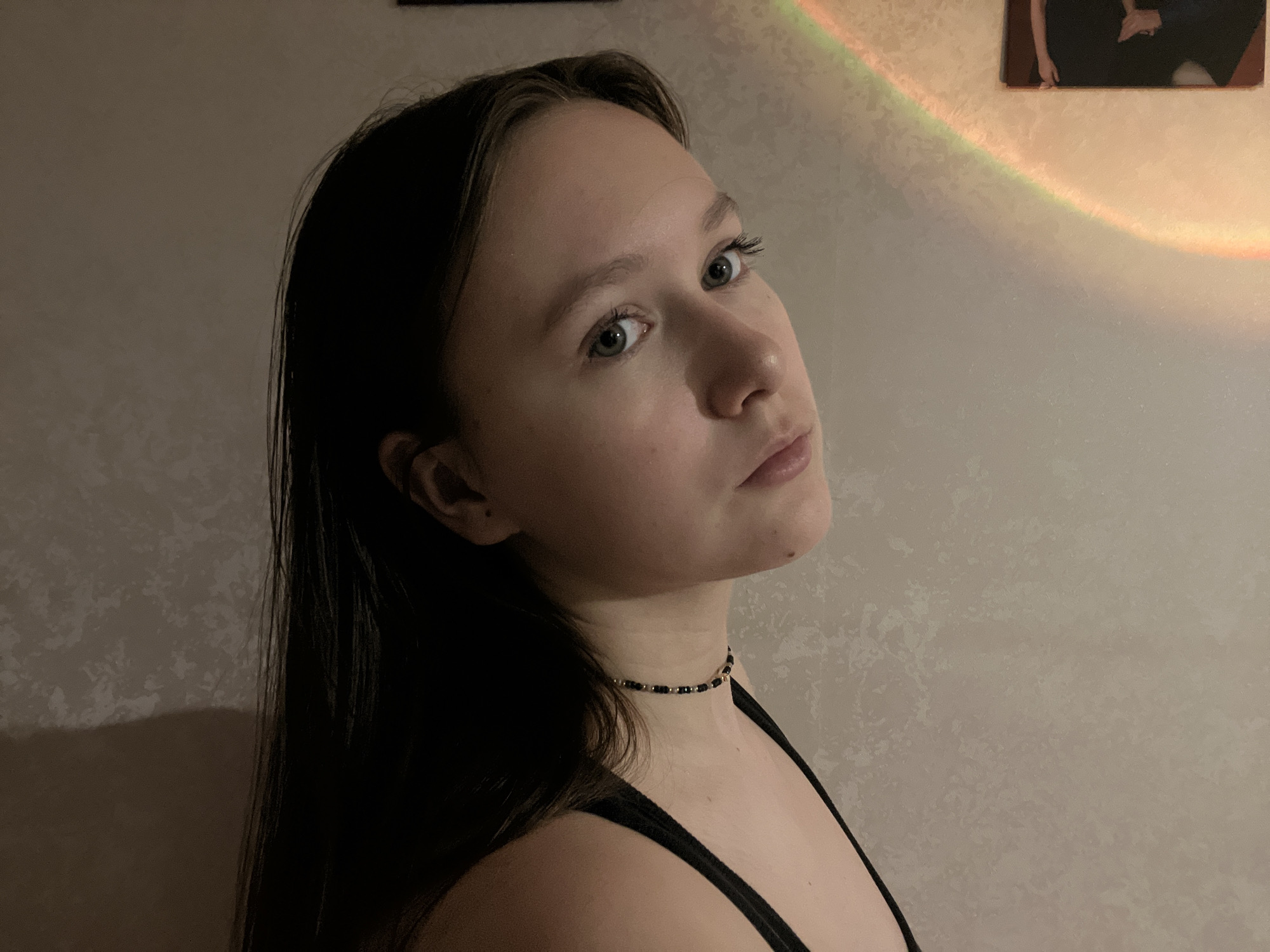 A portrait-style photograph features a young woman, likely taken indoors in Chaikovsky, Russia. She is positioned slightly to the left of the frame, looking upwards and to her right with a neutral to contemplative expression. She has long, dark brown hair, which falls over her shoulders, and light-colored eyes. She is wearing a black spaghetti-strap top and a black beaded choker necklace with metallic accents around her neck.
The background consists of a light-colored wall with a subtle textured pattern. A distinct, diagonal band of rainbow-colored light is visible on the upper right portion of the wall, potentially cast by a suncatcher or a specialized light source. Above this rainbow effect, a small portion of a framed picture or artwork is partially visible, showing what appears to be human figures or hands. The overall lighting is dim, suggesting an evening setting or intentional low-light conditions, creating a quiet and intimate atmosphere. There is no visible text in the image.