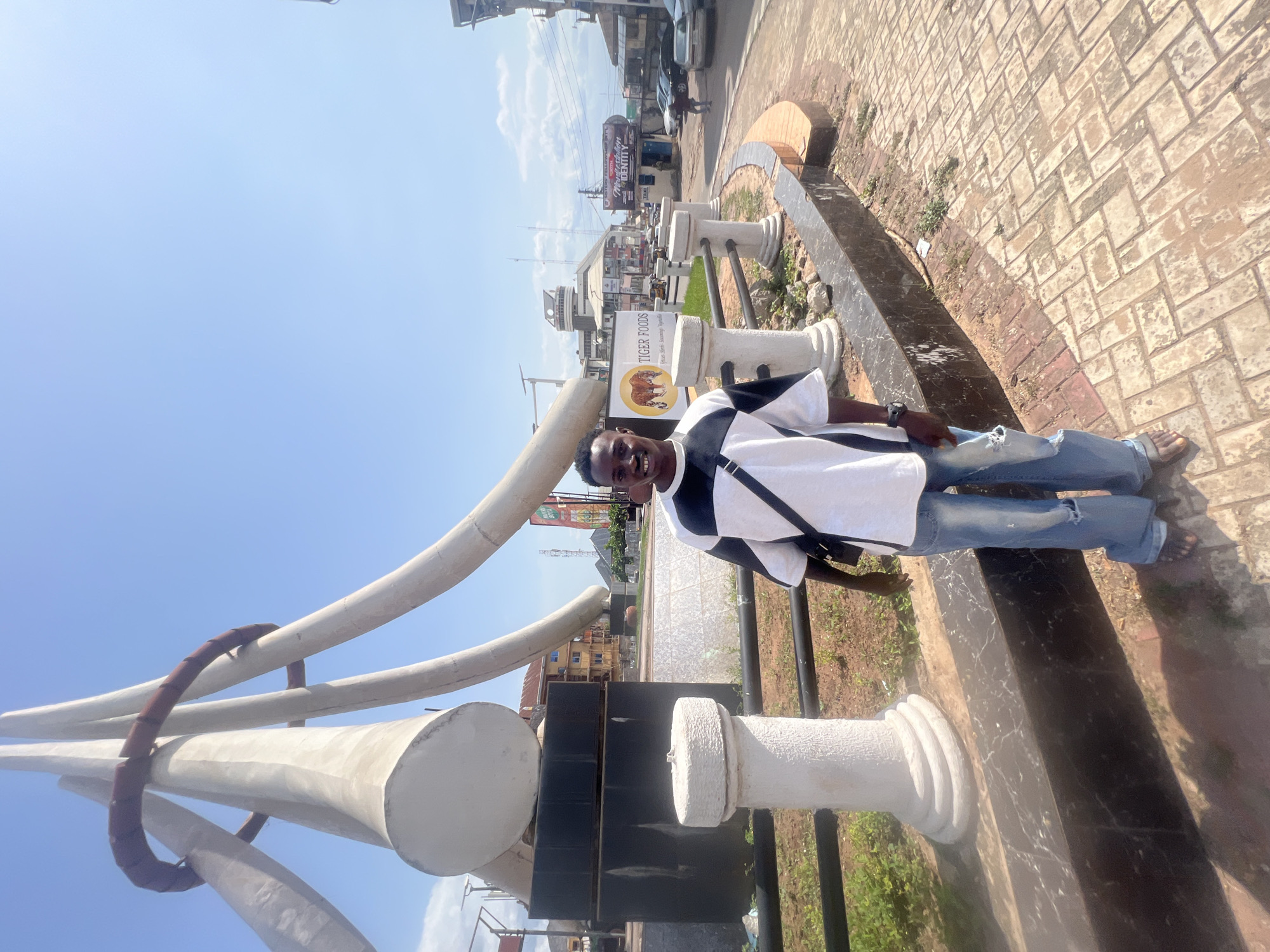 A young man stands smiling brightly in an outdoor setting, likely a public space or street in Onitsha, Nigeria. He is positioned facing the camera, wearing a white short-sleeved shirt with black geometric patterns, blue distressed jeans with a tear on the left knee, and sandals. A dark strap from a bag crosses his chest.
To his right, there are decorative white pillars and a long, dark-colored raised planter or barrier made of what appears to be marble. Further to his left, large, abstract curved white structures, resembling modern art or stylized tusks, extend upwards and outwards against the clear blue sky.
The background reveals a bustling urban street scene under bright daylight, with scattered clouds visible. Several multi-story buildings line the street, hosting various businesses. A prominent sign for "TIGER FOODS" features an illustration of two elephants and the slogan "Nutritious. Sumptuous. Appetizing." Another sign, partially visible further back, advertises "MARATHON DENTAL." The ground is paved with brown interlocking bricks, and there are patches of dry earth and sparse greenery. The strong sunlight casts distinct shadows, suggesting it is midday or early afternoon. The overall impression is one of a cheerful moment captured in a lively city environment.