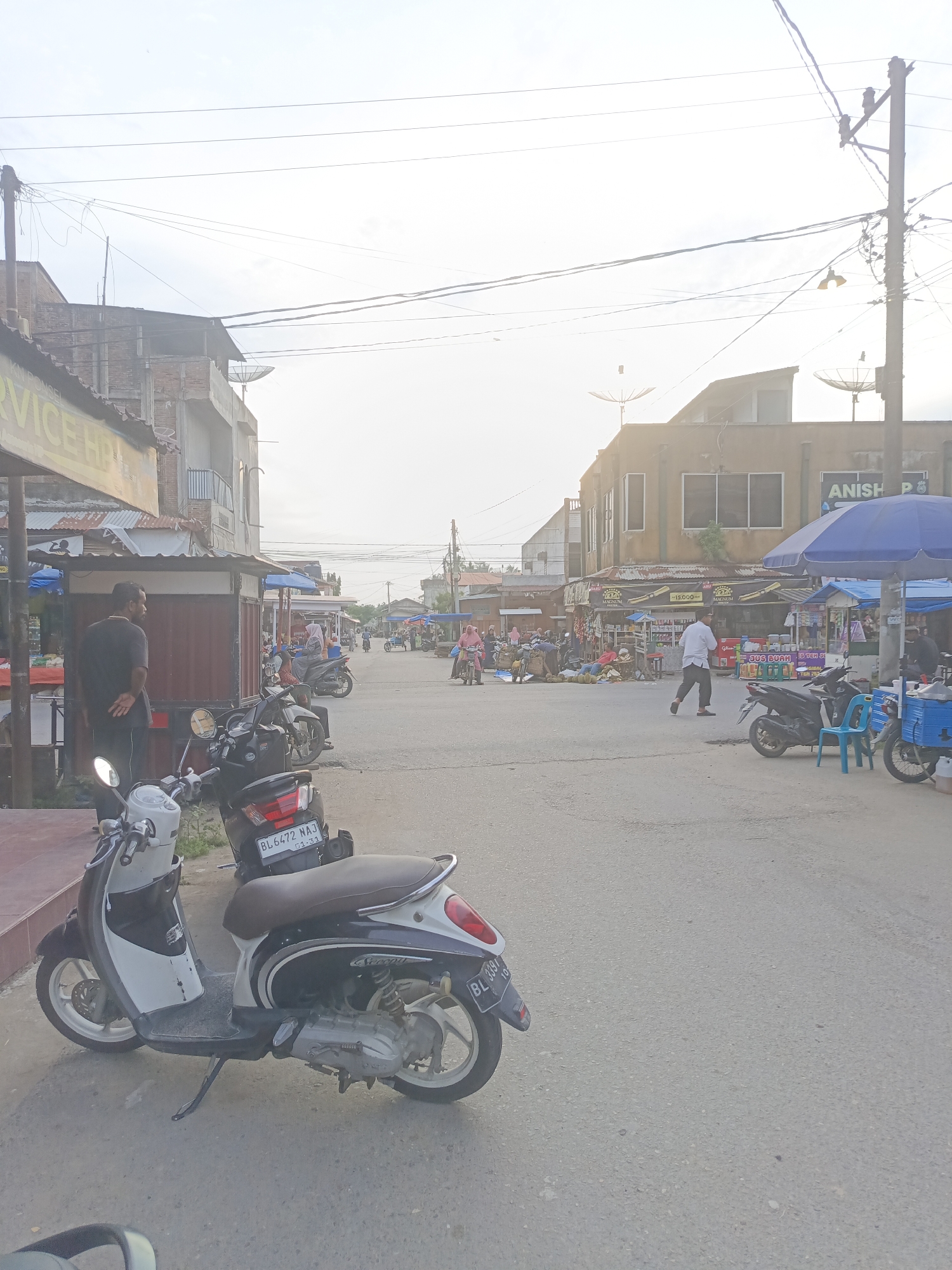 The image depicts a street scene in Lhokseumawe, Indonesia, likely a market area. In the foreground, two scooters are parked. The scooter on the left is white and black with a brown seat, while the scooter on the right is black with a similar brown seat and a license plate reading "BL 6472 NAJ". Further down the street, several people are visible. A man in a black shirt stands near a stall on the left. More stalls line the street, with awnings and products displayed. Several individuals are walking or standing near these stalls. A woman in a pink headscarf rides a motorcycle in the middle ground, and another woman on a motorcycle with a passenger wearing a grey headscarf is seen further back. Buildings with signs, some in Indonesian, are present on both sides of the street. Overhead, power lines crisscross the sky, and a few satellite dishes are mounted on the buildings. The overall lighting suggests it is daytime, possibly late afternoon, with the sun creating a bright, somewhat hazy sky. The ground is paved with asphalt.