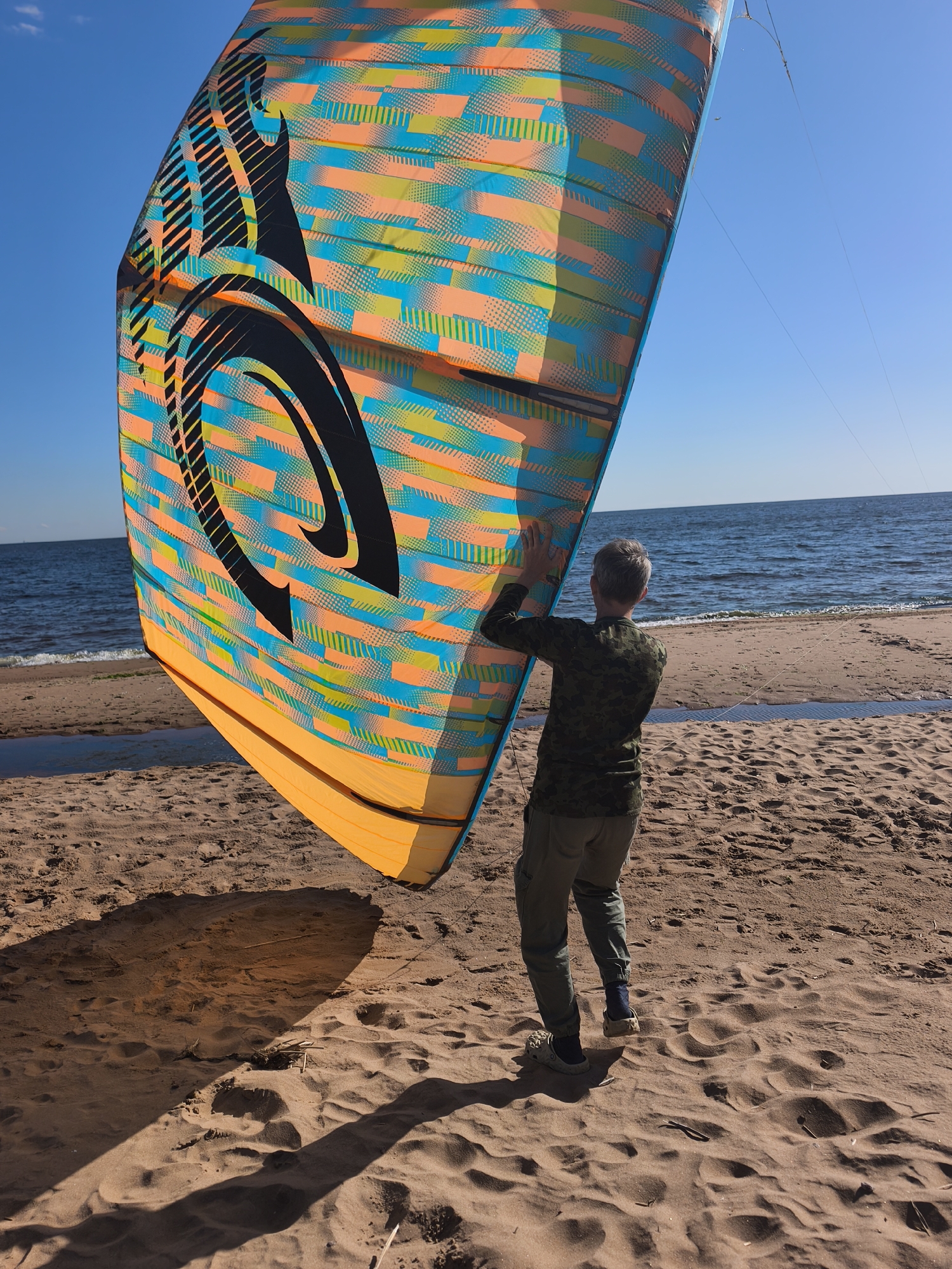 A person wearing a camouflage long-sleeved shirt, olive green pants, and light-colored Crocs stands on a sandy beach, facing away from the camera. They are holding a large, brightly colored kite, which features a black graphic design against a pattern of orange, teal, and lime green rectangles and dots. The kite is positioned at an angle, partially obscuring the sky and the ocean. The person's hands are touching the kite's surface. The sand on the beach shows textures from wind and footprints. In the background, the calm, blue ocean stretches to the horizon under a clear blue sky. The scene appears to be set in the city of Zelenaya Roshcha, Russia, near the coast.
