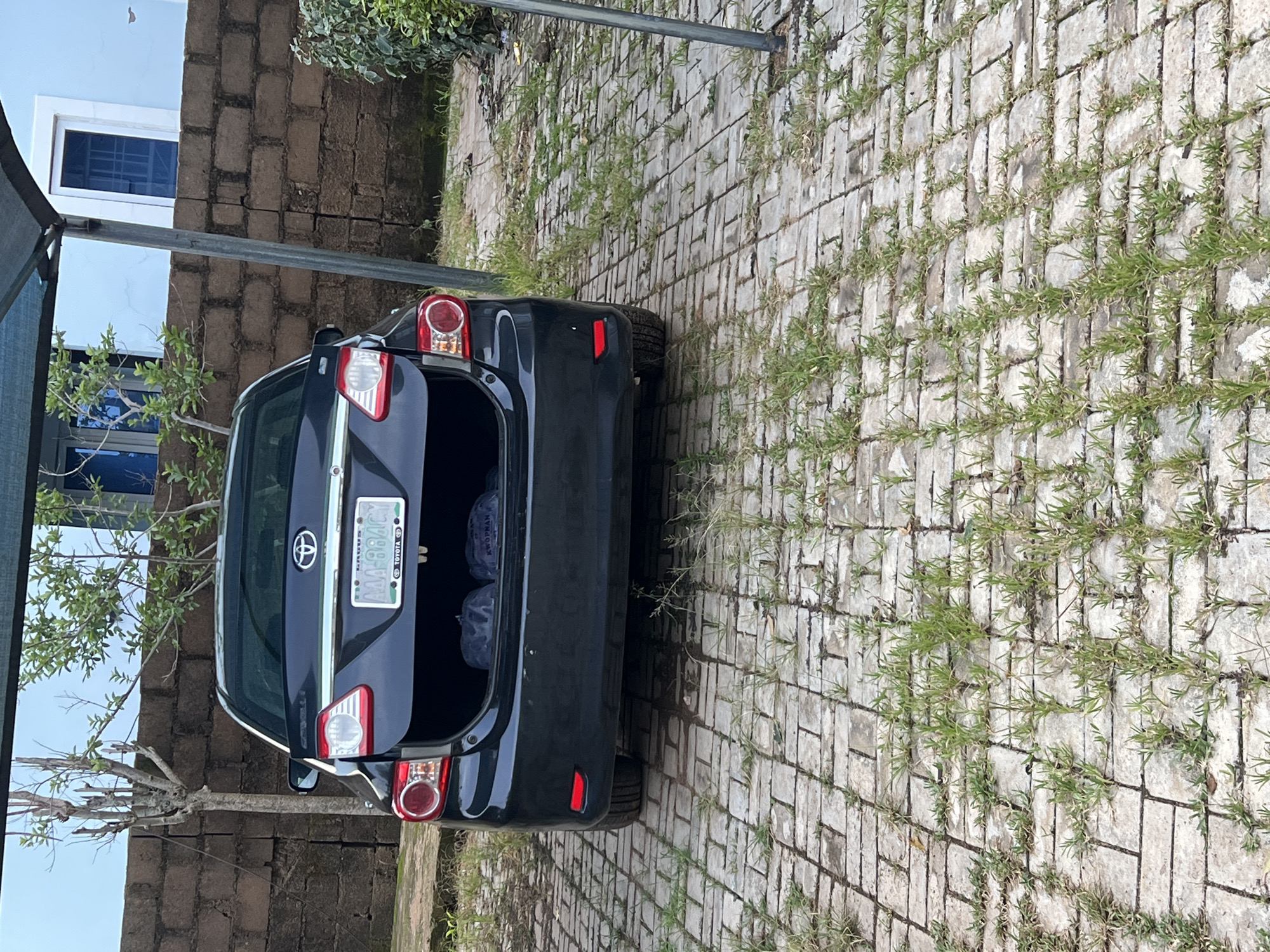 A black 2008-2013 Toyota Corolla Altis sedan is parked on a grey brick paver driveway under a blue awning with a metal support pole. The trunk of the car is open, revealing two dark-colored bags. The car has a license plate that reads "LAGOS AAA-884CY". The car is parked in front of a brick wall and a light blue building. Green vegetation is growing between the bricks of the driveway and also to the right of the car. The scene appears to be in Gura Top, Nigeria, an area known for its residential and commercial developments.