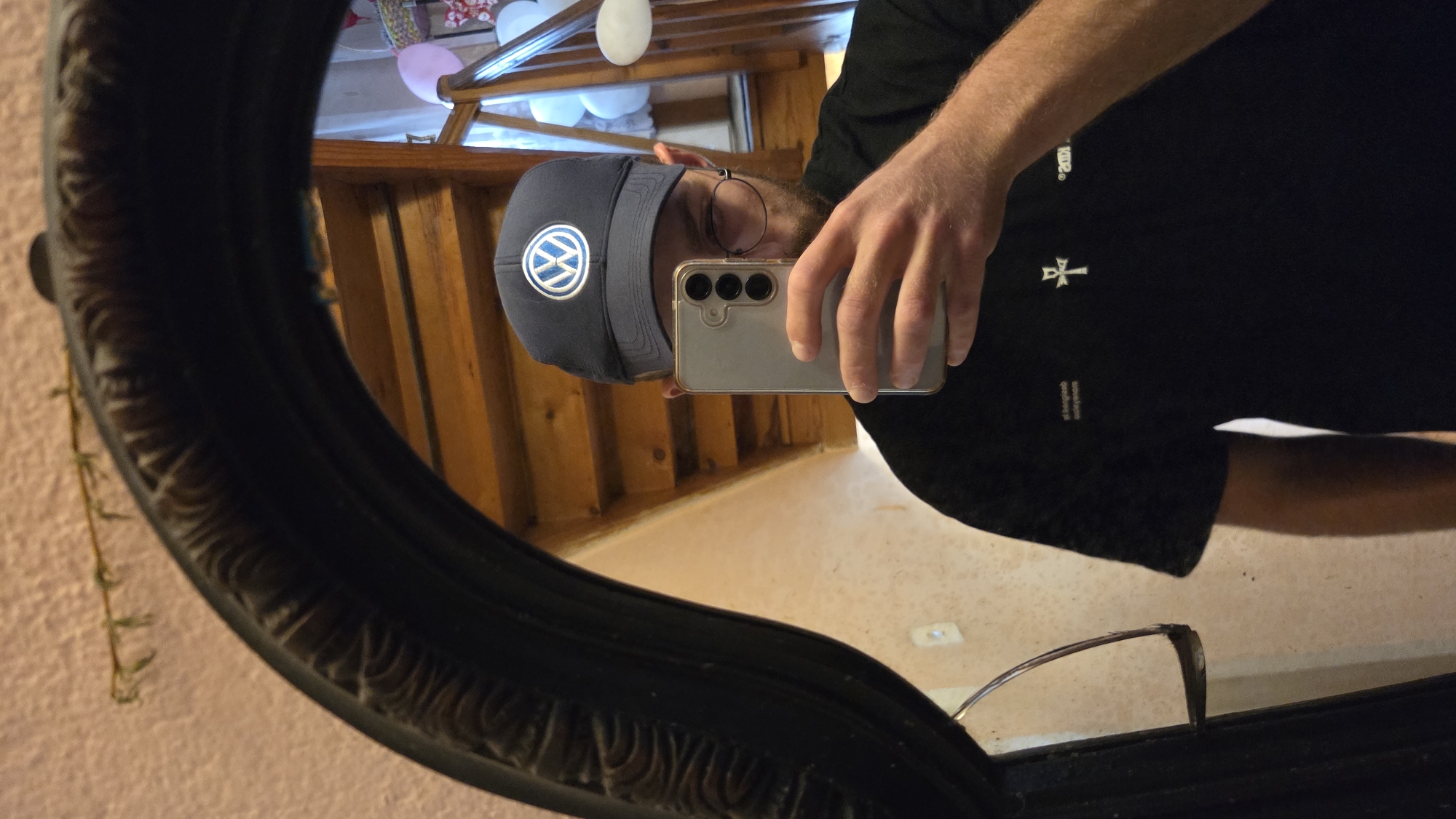 A person is taking a mirror selfie in what appears to be an indoor setting, possibly a home. They are wearing a dark baseball cap with a white Volkswagen logo on the front and a black t-shirt. The t-shirt has some white text and a small white symbol on it, resembling an ankh. The person is holding a silver smartphone with multiple cameras in their right hand, angled to capture the reflection in the mirror. They are also wearing round-rimmed glasses.
The mirror itself is oval-shaped with a dark, ornate frame, and it reflects a wooden staircase in the background. The walls are textured and light-colored. Some pink and white balloons are visible in the reflection on the right side, suggesting a celebratory occasion. The lighting appears to be from an overhead source, creating some shadows. The overall scene is casual and personal.