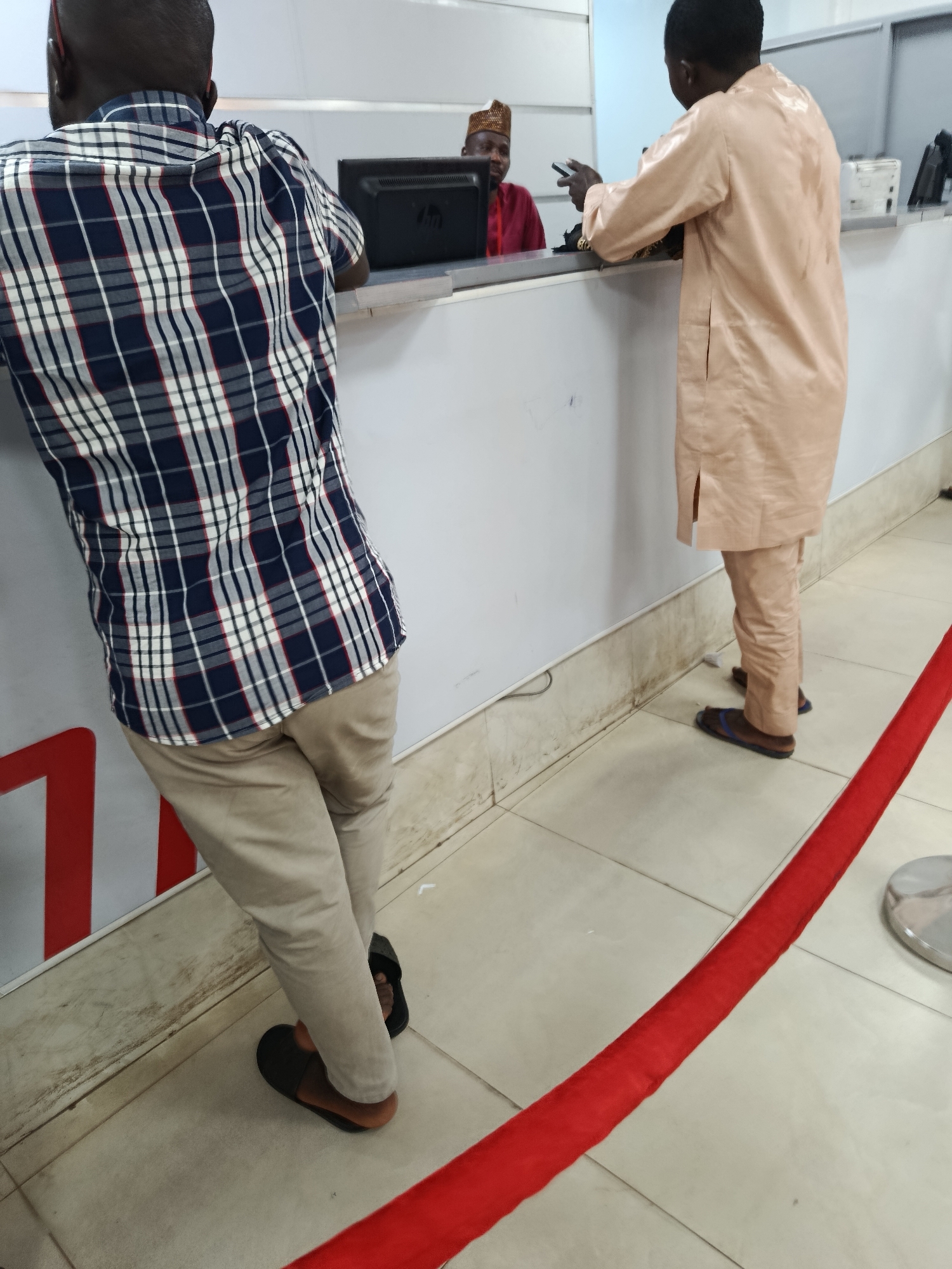 Two men are standing at a counter in what appears to be a public service setting, possibly a bank or government office, given the presence of a counter and computer equipment. One man, seen from the back, is wearing a blue, white, and red plaid shirt and tan pants. He is leaning on the counter. The other man, also viewed from the back, is wearing a light peach-colored traditional outfit consisting of a long tunic and matching pants. He is interacting with someone behind the counter.
In the background, a third person, wearing a red shirt and a patterned cap, is visible behind the counter, suggesting they are an employee or official. A computer monitor is on the counter. A red velvet rope is laid out on the tiled floor, directing a queue. The scene is indoors, with neutral-colored walls and floor tiles. There is no indication of the time of day or weather. The overall atmosphere appears to be one of routine service interaction. No discernible text is visible in the image. The location context of Jos, Nigeria, suggests a common urban environment.