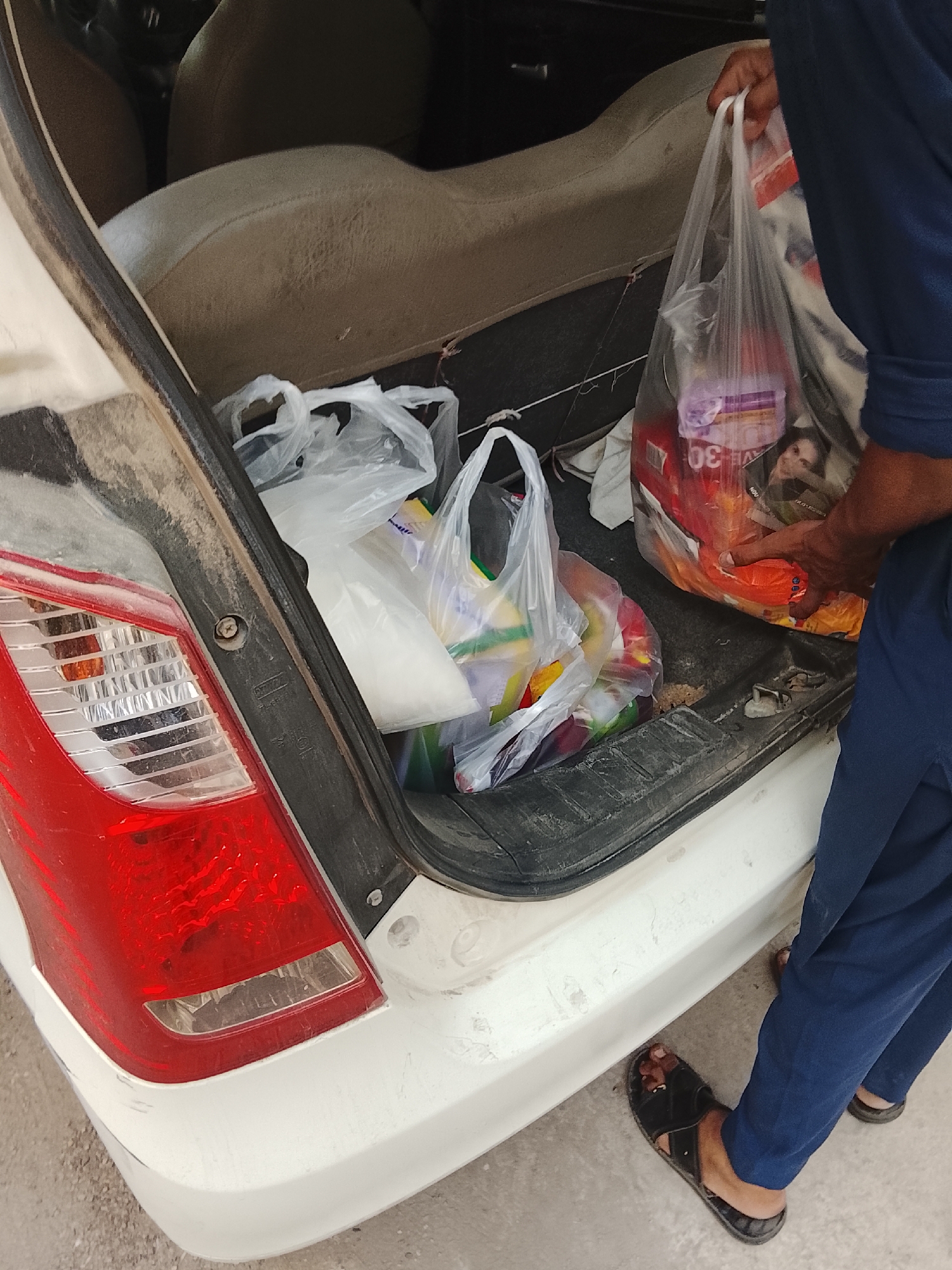 A man is loading grocery bags into the trunk of a dirty white car. The car's taillight is visible on the left. The man is wearing blue clothing and black sandals. The trunk contains several plastic bags filled with groceries. The photo was taken in Gulgasht Colony, Pakistan.