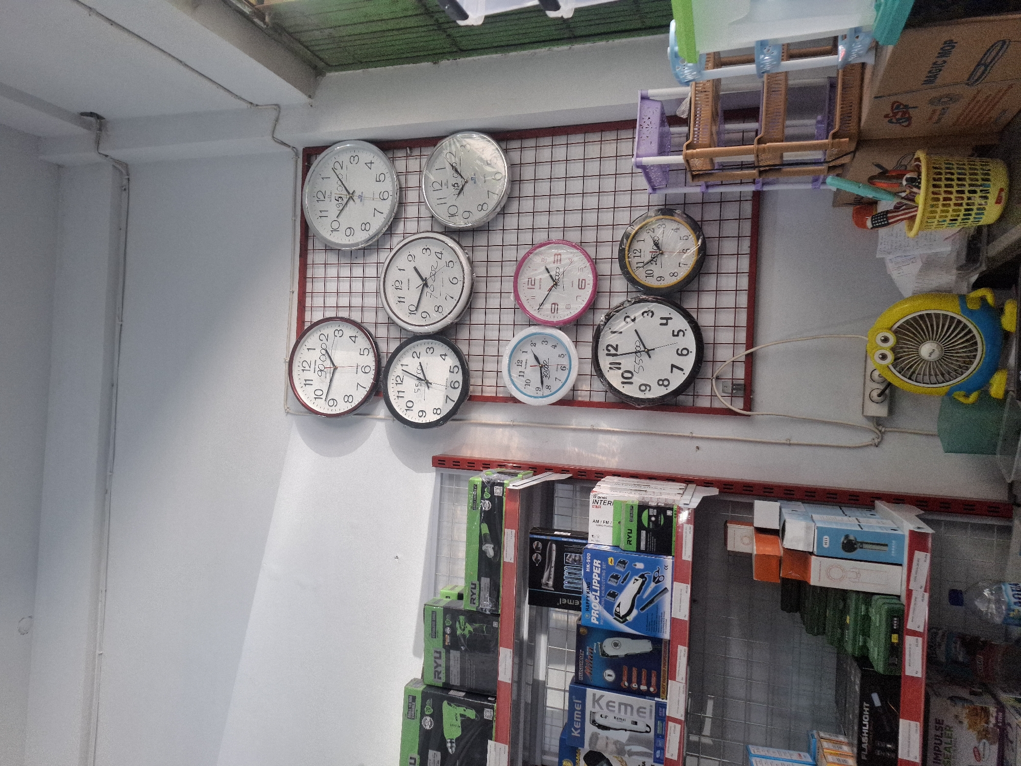 The image depicts a retail store interior in Sumberwudi, Indonesia. The scene is dominated by shelves stocked with various products, primarily electronics and household items. On the right wall, a grid display features numerous wall clocks of different sizes and designs. Among the products on the shelves are power tools, hair clippers, flashlights, and impulse sealers. A small yellow fan with a cartoonish design is visible on a lower shelf, near a yellow basket filled with pens and other small items. A bottle of water and several boxed products are also present. The environment appears to be a well-lit store, with the main light source appearing to be overhead.