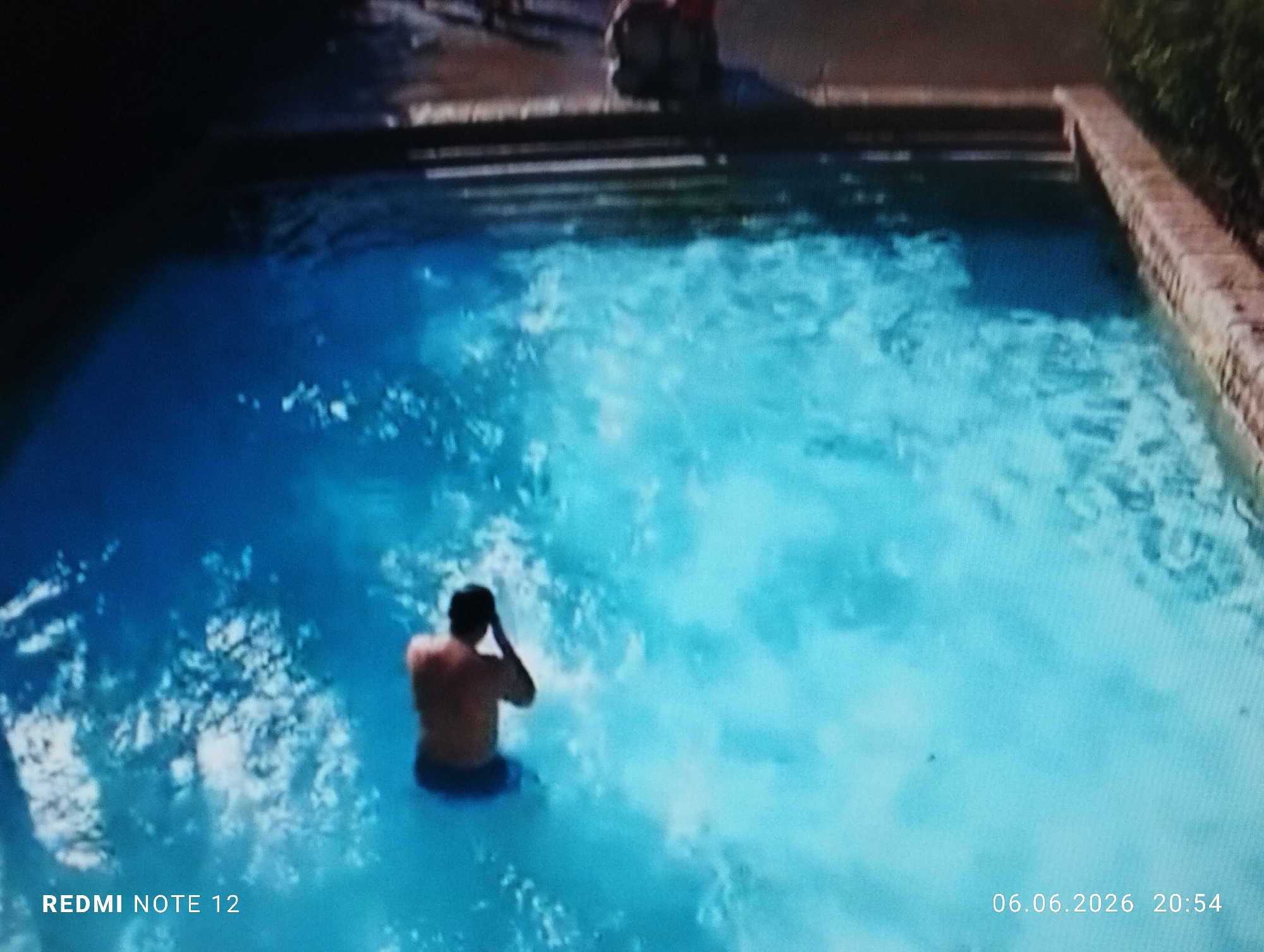 An overhead, slightly blurred image captures an outdoor swimming pool scene. A single male individual, seen from behind, is partially submerged in the light blue water, wearing dark swim trunks and bare-chested. His hands are raised to his head. Significant splashing and ripples emanate from his position in the water, indicating recent movement. The pool perimeter features a reddish-brown coping, and steps descending into the water are visible at the far end. Beyond the pool, a paved area is present, with highly blurred figures discernible in the distance. The right side of the pool is bordered by a stone wall with green foliage. Text overlays in the lower left corner identify the device as "REDMI NOTE 12," and in the lower right corner, the date and time are displayed as "06.06.2026 20:54." The precise location in the city of Burgas, Bulgaria, cannot be ascertained from the visual information provided in the image.