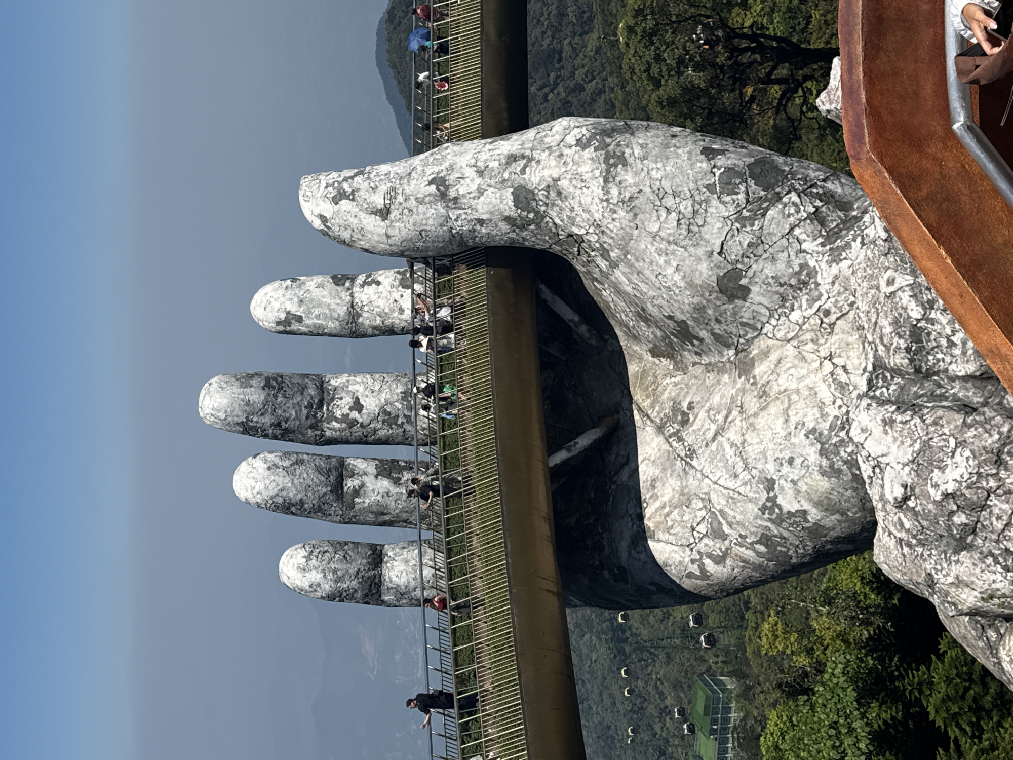 The image depicts the Golden Bridge in Bà Nà Hills, Đà Nẵng, Vietnam. The bridge is supported by two giant, stone-like hands. People are walking on the bridge. Lush green foliage covers the surrounding hillsides. In the distance, a clear sky transitions into a hazy grey. A cable car line is visible in the background.