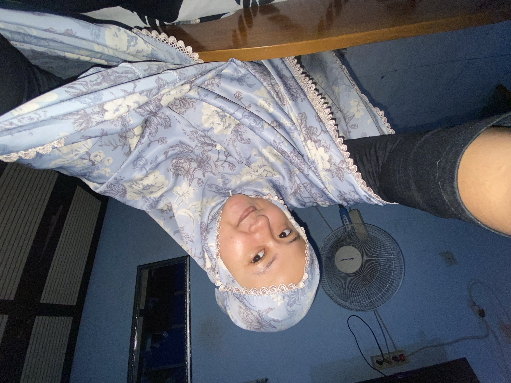 A selfie is taken by a woman wearing a light blue floral hijab and black clothing. She is looking at the camera with a slight smile, and her face is illuminated. In the background, a round white fan is visible on a blue wall. Electrical outlets and cords are also present on the wall. To the right, a mirrored surface with a black frame reflects a dimly lit area. The scene appears to be indoors, possibly a bedroom or living space in Gelam, Indonesia, suggested by the domestic setting and typical household items. The overall lighting is dim, with a slight blue cast from the walls.