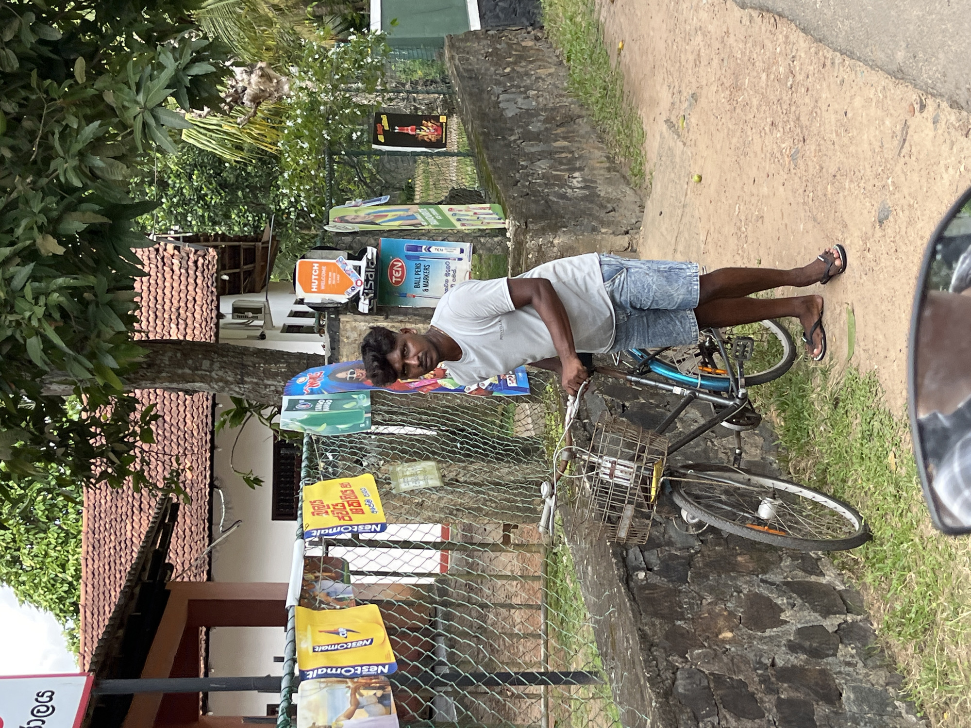 A man stands next to a bicycle on a dirt road in Kabalana, Sri Lanka. He is wearing a light gray t-shirt and blue denim shorts, with dark skin and dark hair. The bicycle is dark-colored with a wire basket on the front. Behind the man and the bicycle is a chain-link fence with several colorful advertisements attached to it. The advertisements are for various products, including "Nestomalt," "HUTCH," and "TEN BALL PENS & MARKERS." Beyond the fence, a building with a red tile roof and white walls is visible. Green trees and foliage surround the area, indicating a tropical environment. The ground is a mix of dirt and patchy grass, with a concrete wall bordering the road. The lighting suggests it is daytime.