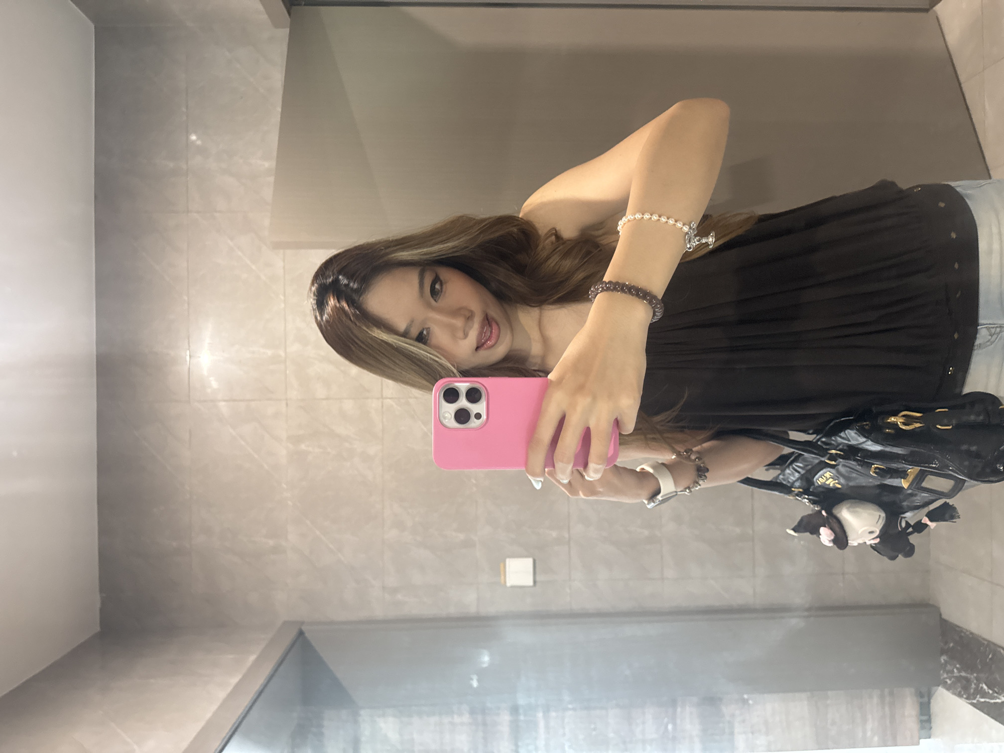 A woman is taking a selfie in a bathroom with light gray walls. She is holding a pink phone in her left hand. She is wearing a black strapless top, a white bracelet, and a dark beaded bracelet on her left wrist. She has long brown hair with blonde highlights. A shiny black handbag with a white plush toy attached is visible to the right of the woman. The location appears to be inside a modern bathroom, based on the tile pattern and design. It is not possible to verify whether the image was taken in Nan'an District, China.