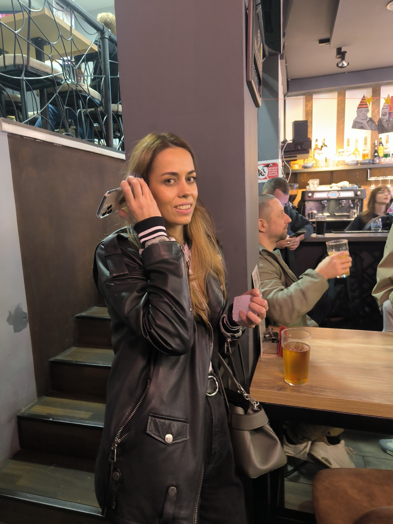An indoor scene in a bar in Bar, Montenegro, is captured from a slightly elevated perspective. In the foreground, a female with long, light brown hair, wearing a black leather jacket and a striped long-sleeved top, stands smiling at the camera. She holds a clear-cased smartphone to her right ear with her right hand and a small white card-like object in her left hand. A grey handbag with a chain strap hangs from her left shoulder. To her immediate right, a wooden table holds a half-full glass of amber liquid, presumably beer.
Behind the female, to the left, a flight of wooden stairs descends into a darker area. Above the stairs, an upper level is visible through a black metal railing with a web-like pattern, showing tables and chairs occupied by patrons.
To the right of the foreground subject, the bar area is visible. A man in an olive green jacket sits at a wooden table, holding a glass of amber liquid, facing left. Behind him, another man in a dark shirt leans against the bar counter, looking at a smartphone. Further right, a woman is partially visible sitting at the bar. The bar counter features bottles of alcohol, an espresso machine, and glassware. Above the bar, two decorative figures (one with a red party hat, the other with face paint) are displayed on the wall, traversed by a thin red horizontal line. A red sign with white Cyrillic text, possibly indicating an age restriction, is partially visible on a grey pillar. The overall lighting is warm, particularly around the bar area.