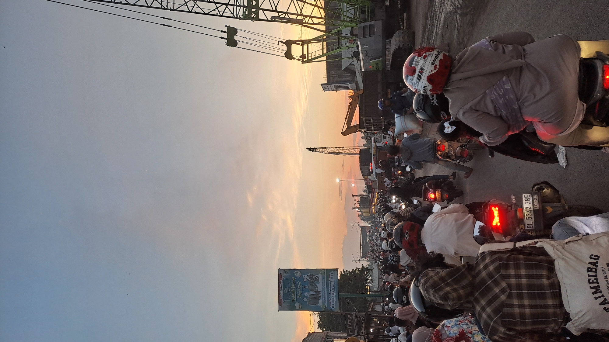 The image depicts a busy street scene with heavy traffic congestion, likely during sunset in Tingkeummanyang, Indonesia. Numerous motorcycles, many carrying multiple passengers, fill the road. People are wearing helmets, and some are dressed in casual clothing. In the background, large construction equipment, including cranes and excavators, are visible, suggesting ongoing development or infrastructure work. A large billboard advertising an event or product stands on the left side of the street. The sky is illuminated with warm hues of orange and yellow, characteristic of a sunset. The overall atmosphere is one of a bustling urban environment experiencing a traffic slowdown.