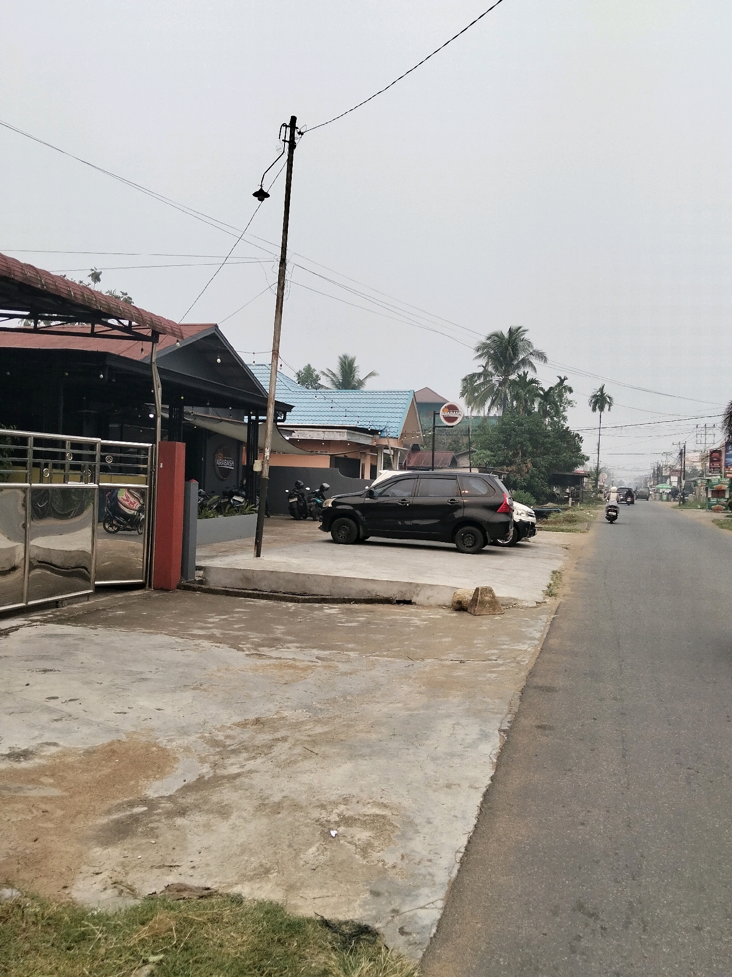 In Arang Limbung, Indonesia, this image captures a roadside scene on a cloudy day. A black SUV is parked on a concrete lot adjacent to a road. To the left of the SUV is a building with a red-tiled roof, partially obscured by a metal gate. A sign with the word "ARABASH" is visible on the building. Several motorcycles are parked in front of the building. The road is paved, and a few other vehicles, including motorcycles, are visible in the distance. Palm trees and other vegetation dot the landscape. The sky is overcast, suggesting a hazy or misty atmosphere. The image is taken from a low-angle perspective, facing the road and the buildings on the left.