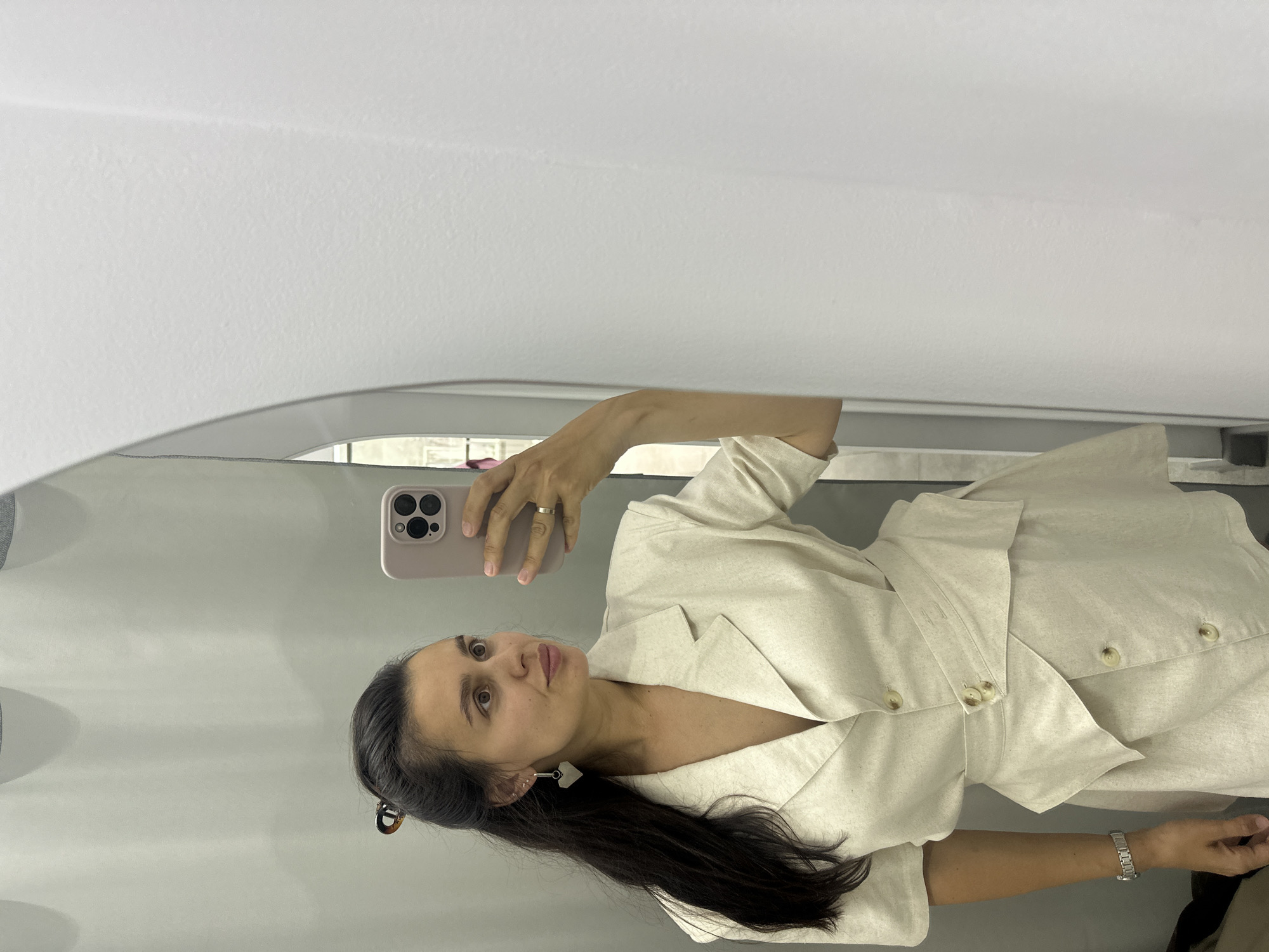 In Chaikovsky, Russia, a woman is captured in a mirror selfie within a changing room. She is wearing a light-colored, short-sleeved blazer dress with a cinched waist. Her hair is dark and styled in a ponytail, adorned with a tortoiseshell clip. She is accessorized with a heart-shaped earring and a watch. The background consists of a grey curtain and a white wall, indicative of a retail dressing area.