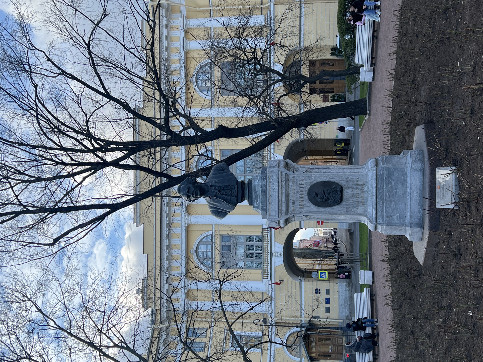 The image shows a monument to Mikhail Lomonosov, a Russian polymath, in Saint Petersburg, Russia. The monument, featuring a bust of Lomonosov, stands in front of the Kunstkamera, a museum of anthropology and ethnography. Bare tree branches frame the monument and the yellow facade of the Kunstkamera in the background. Pedestrians walk through an archway in the building. People sit on benches near the monument. The sky is visible through the tree branches.