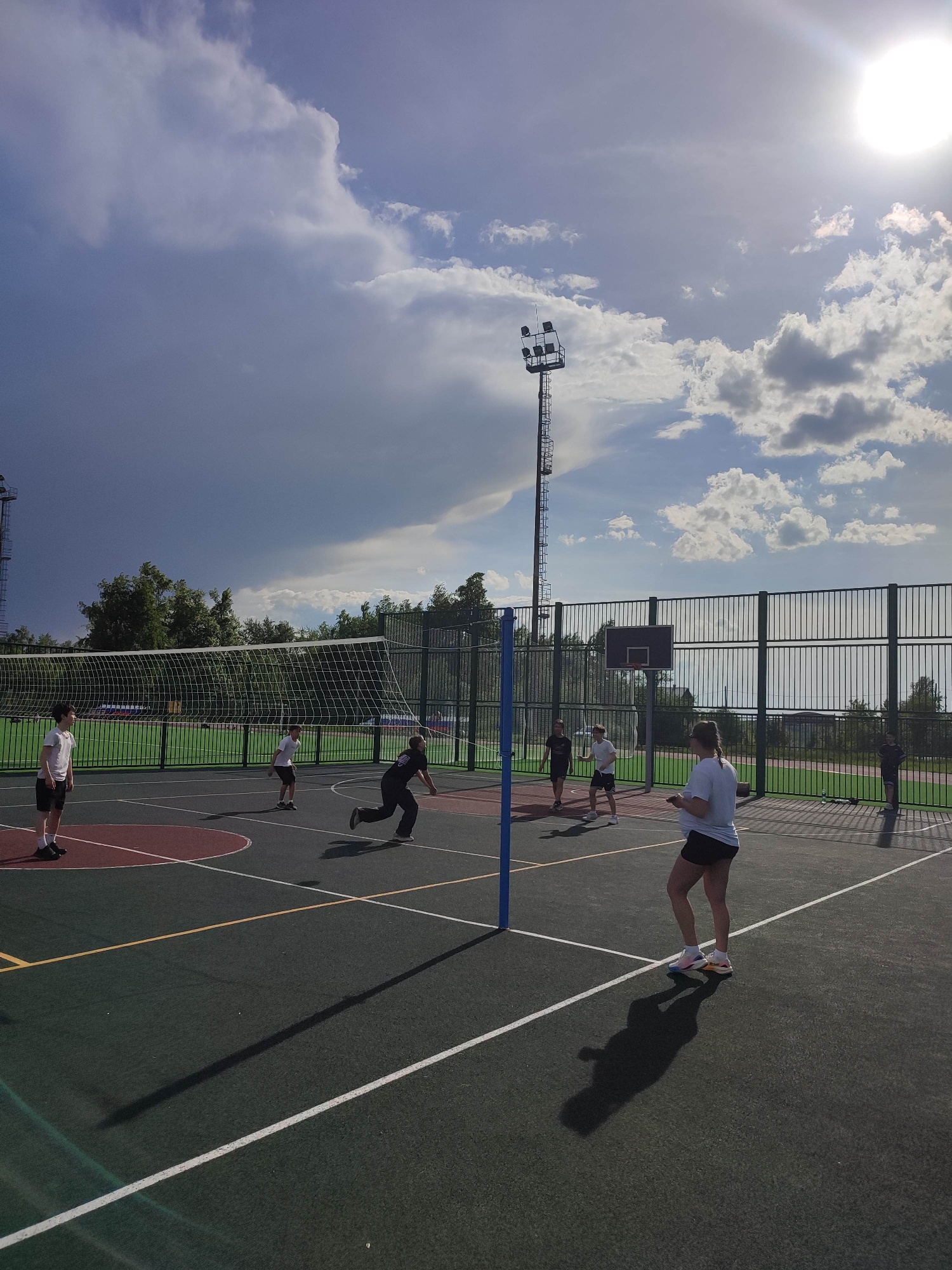 The image shows a group of people playing volleyball on an outdoor court. The court has a green surface with white lines marking the boundaries. A volleyball net is set up in the middle of the court. A fence surrounds the court, and there are trees in the background. A tall light post stands near the fence. The sky is partly cloudy, with a mix of dark and light clouds. The sun is visible, shining brightly. The location is Unknown, Unknown.