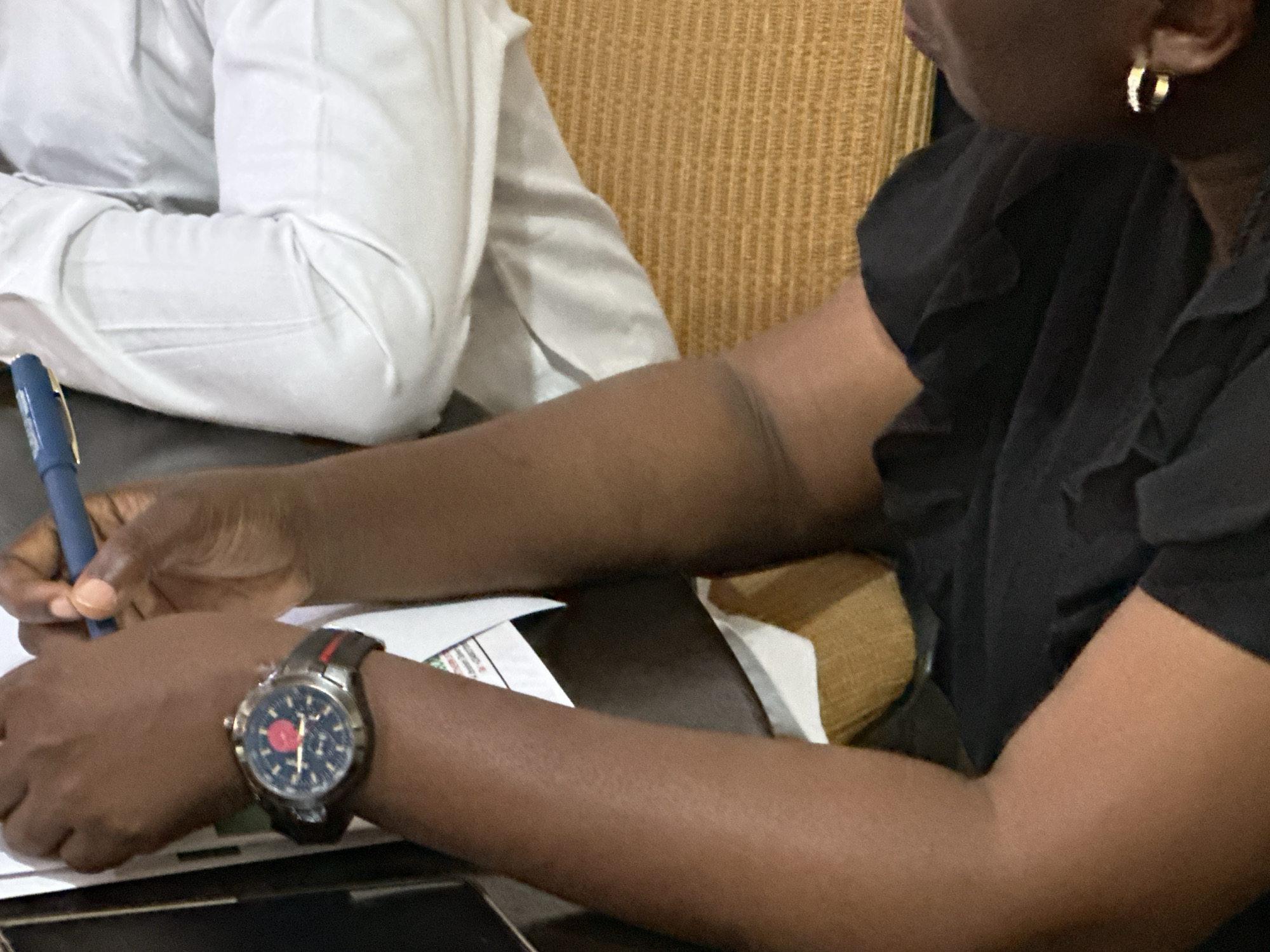 The image shows a close-up of two individuals sitting at a table, with a focus on their arms and hands. The person on the left is wearing a white collared shirt, and their left arm is visible. A blue pen is held in their right hand, which is positioned over some papers on the table. On their wrist, they are wearing a wristwatch with a dark band featuring a red stripe, and a dark face with a red circular accent. The person on the right is wearing a black top with ruffled sleeves and a gold hoop earring. Their right arm is also visible, and they are seated next to the other person. The background consists of a textured, yellowish-brown surface, likely upholstery, and a white surface that could be part of a wall or another object. The scene appears to be indoors, possibly in an office or meeting room, given the presence of papers and the professional attire. The lighting suggests it is daytime. The overall mood is neutral, focusing on the actions of writing and the objects present. There is no visible text in the image.