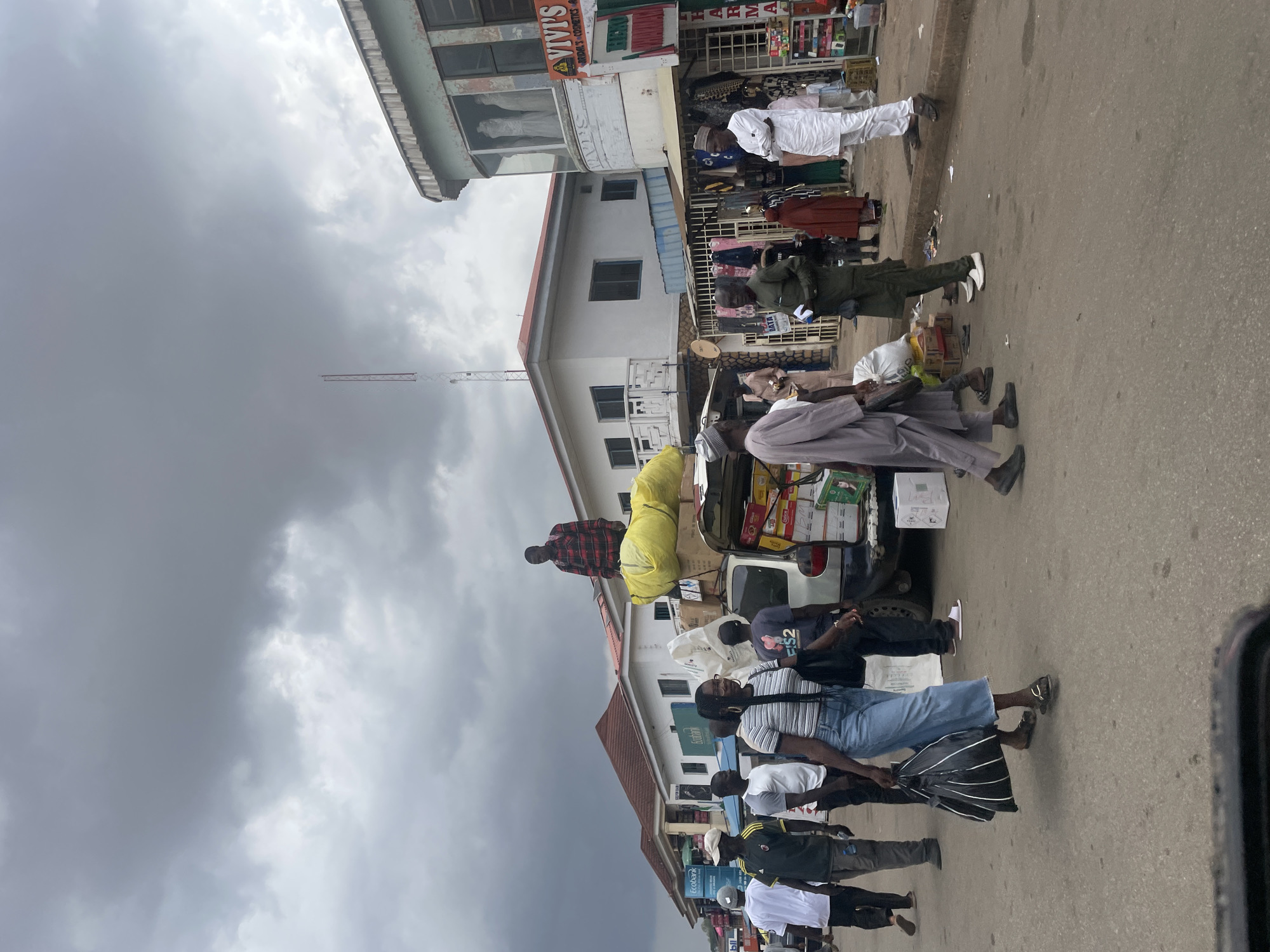The image depicts a street scene, possibly a market area, in Jos, Nigeria. A line of people are walking along the side of the street next to cars parked on the shoulder of the road. The people wear casual clothing. One man is wearing a military style uniform. A man standing in the back of a car appears to be loading it with yellow bags. In the background, there are two-story buildings with storefronts. A tall antenna extends into a cloudy sky.