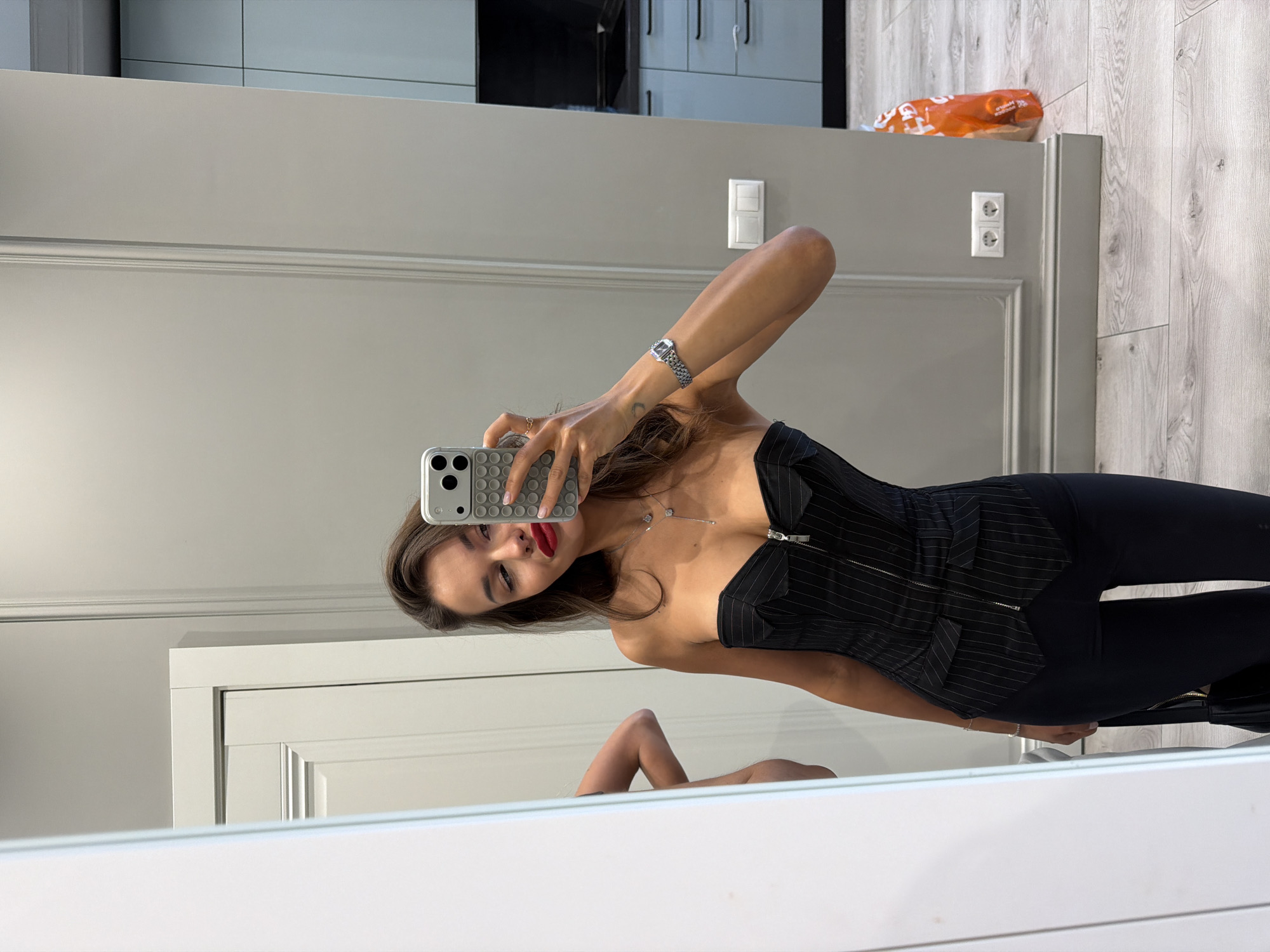 A young adult woman stands in a modern apartment in Khimki, Russia, taking a mirror selfie with a smartphone. She has long brown hair, wears bright red lipstick, and is dressed in a black pinstriped strapless corset top with a front zipper, and dark pants. Her right hand, with a ring on her index finger, holds a grey smartphone featuring a textured, possibly 'pop-it' style, case and multiple camera lenses, positioned to capture her reflection. A silver watch with a mesh band is visible on her left wrist, and a delicate silver necklace with two square pendants adorns her neck. A partial reflection of a bare arm and back, possibly her own, is visible in the mirror to the left. The apartment features light grey walls with white architectural molding and baseboards. White electrical outlets and light switches are mounted on the wall. In the background, a kitchen area with light blue-grey base cabinets and a dark countertop and stovetop is visible. The flooring consists of light-colored wood laminate. An orange plastic bag rests on the floor near the kitchen area, and a black handbag is partially visible, held in her left hand near her hip.