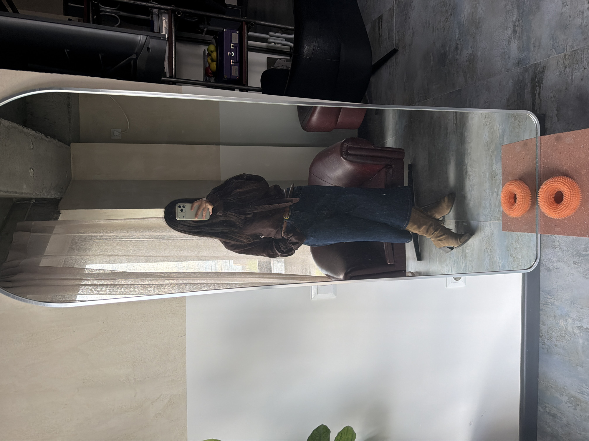 This full-length mirror selfie captures an individual dressed in a dark brown top or jacket, dark blue skirt or wide-leg trousers, and light brown ankle boots, taking a photo with a white/light-colored smartphone that obscures their face. A brown belt is visible around their waist, and they have long, dark hair.
The setting appears to be an indoor space, likely a living room or hallway in Yerevan, Armenia. The room features light-colored walls and a textured gray tiled floor, resembling concrete. Natural light enters from a window on the left, partially covered by a sheer, light-colored curtain.
Reflected in the mirror are several pieces of furniture and decor: two leather armchairs, one dark reddish-brown and another darker, are positioned behind the person. Further back, a dark shelving unit holds various items, including a purple box and a bowl of fruit (apples or pears). To the right of the mirror, on a terracotta-colored surface, two distinct round, orange, spiky or textured decorative objects are visible.
The overall aesthetic is modern and minimalist. The image appears to be taken during the daytime due to the natural light. No visible text is clearly discernible, apart from a potential logo on the phone case. The image is presented rotated 90 degrees clockwise.