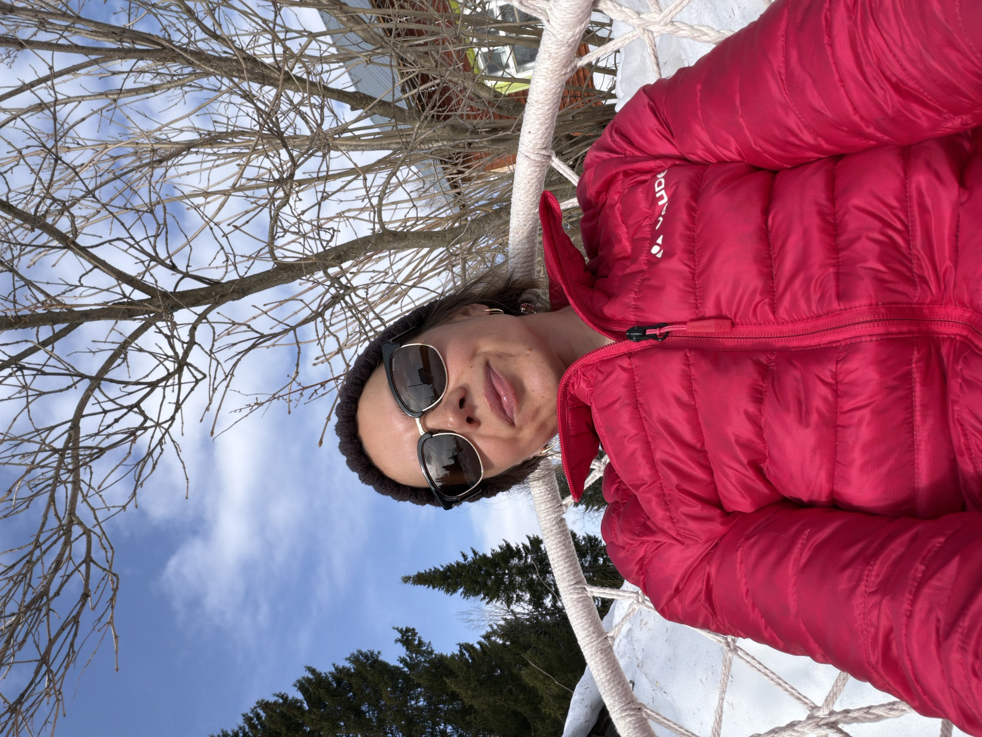 An individual is depicted in a selfie-style portrait, seated outdoors in a snowy environment within the city of Верх-Язьва, Russia. The subject, an adult female, is positioned centrally, looking directly at the camera with a subtle smile. She is wearing dark sunglasses with reflective lenses, a dark knit beanie, and a bright pink VAUDE-branded puffer jacket with a visible black zipper pull. She is seated in a white, woven rope hammock chair, elements of which are visible to the left and right of her body. The ground beneath and around her is covered in white snow. The background features several tall, green coniferous trees on the left. In the center and right background, numerous deciduous trees with bare branches are visible, partially obscuring a structure that appears to be a building with an orange-red facade, a light green window frame, and a grey corrugated roof. The sky is predominantly clear blue with scattered white clouds, and the scene is illuminated by bright natural sunlight.