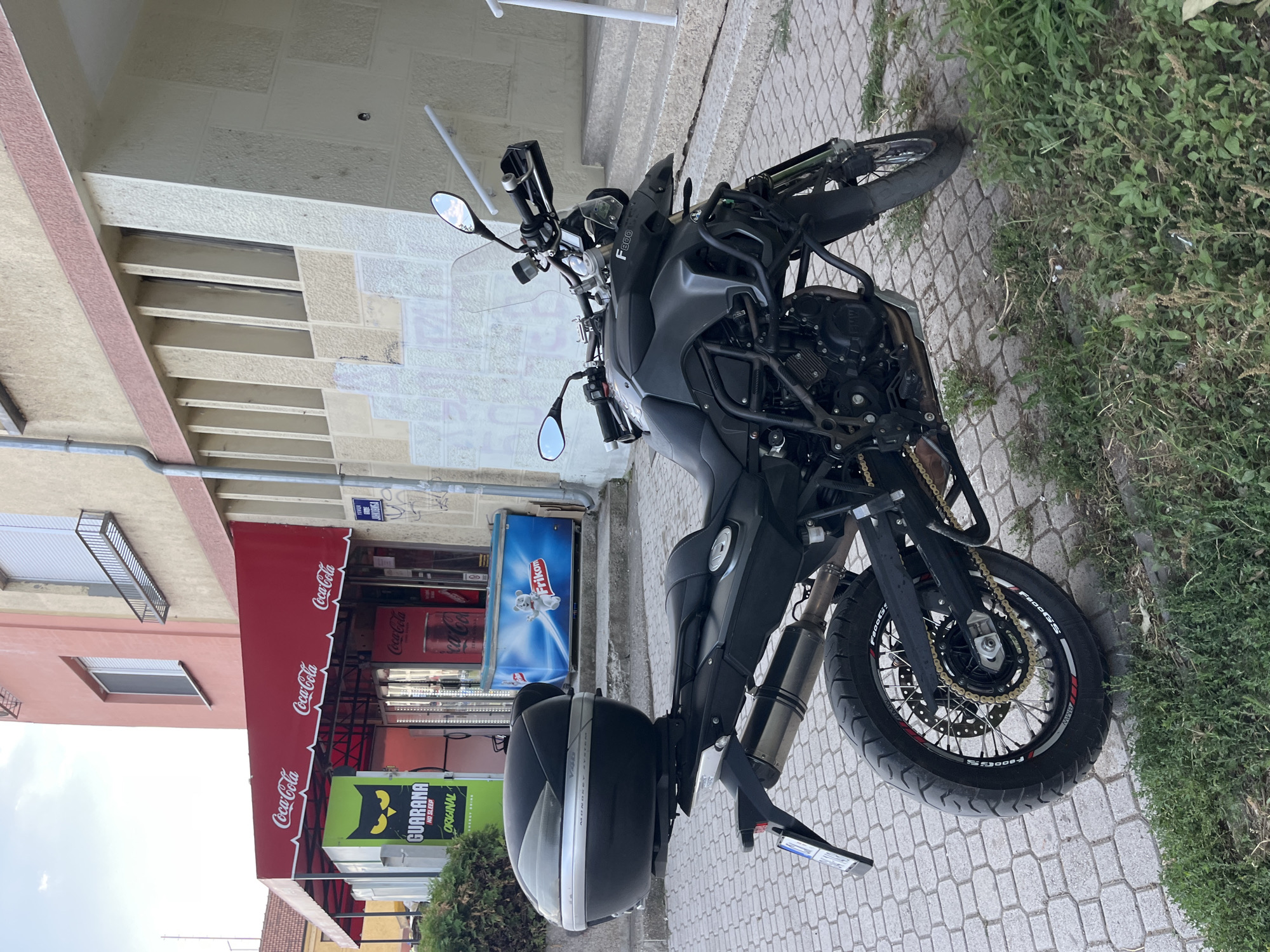 A black BMW F800GS motorcycle is parked on a cobblestone sidewalk in front of a building with a red awning. The awning has "Coca-Cola" logos. To the left of the awning, there is a green sign with a black cat silhouette and "Guarana Original" text. To the right of the awning, there is a blue Frikko branded freezer. The motorcycle has a black top case and a metallic exhaust pipe. The background shows the facade of the building with windows and decorative elements. A street sign that reads "Pula-Danilovgrad" is visible on the right side of the image. The environment appears to be an urban setting in Cacak, Serbia.