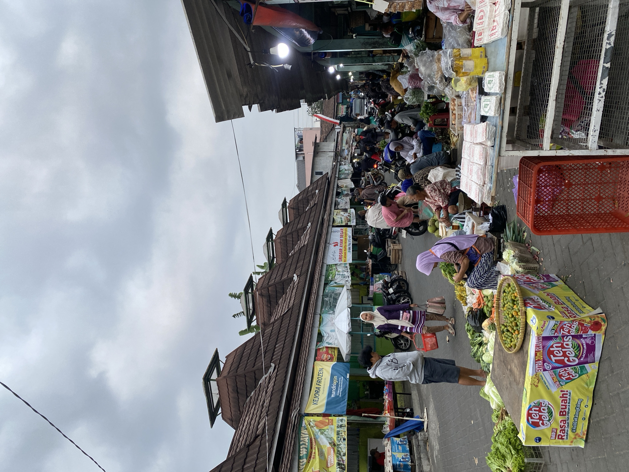 The image depicts a busy outdoor market scene in Ambarketawang, Indonesia. The market is lined with stalls under a covered walkway with traditional tiled roofs. Banners advertising various products are hung along the front of the stalls, including "KEJORA FROZEN" and "GROSIR AYAM SEGAR". People are engaged in commerce, with some vendors selling vegetables and other goods displayed on tables and on the ground. Several individuals are seen browsing or purchasing items. Motorcycles are parked and present throughout the market, indicating common modes of transportation. The overall atmosphere is that of a lively local marketplace with goods and people in abundance. The sky is overcast, suggesting a cloudy day.