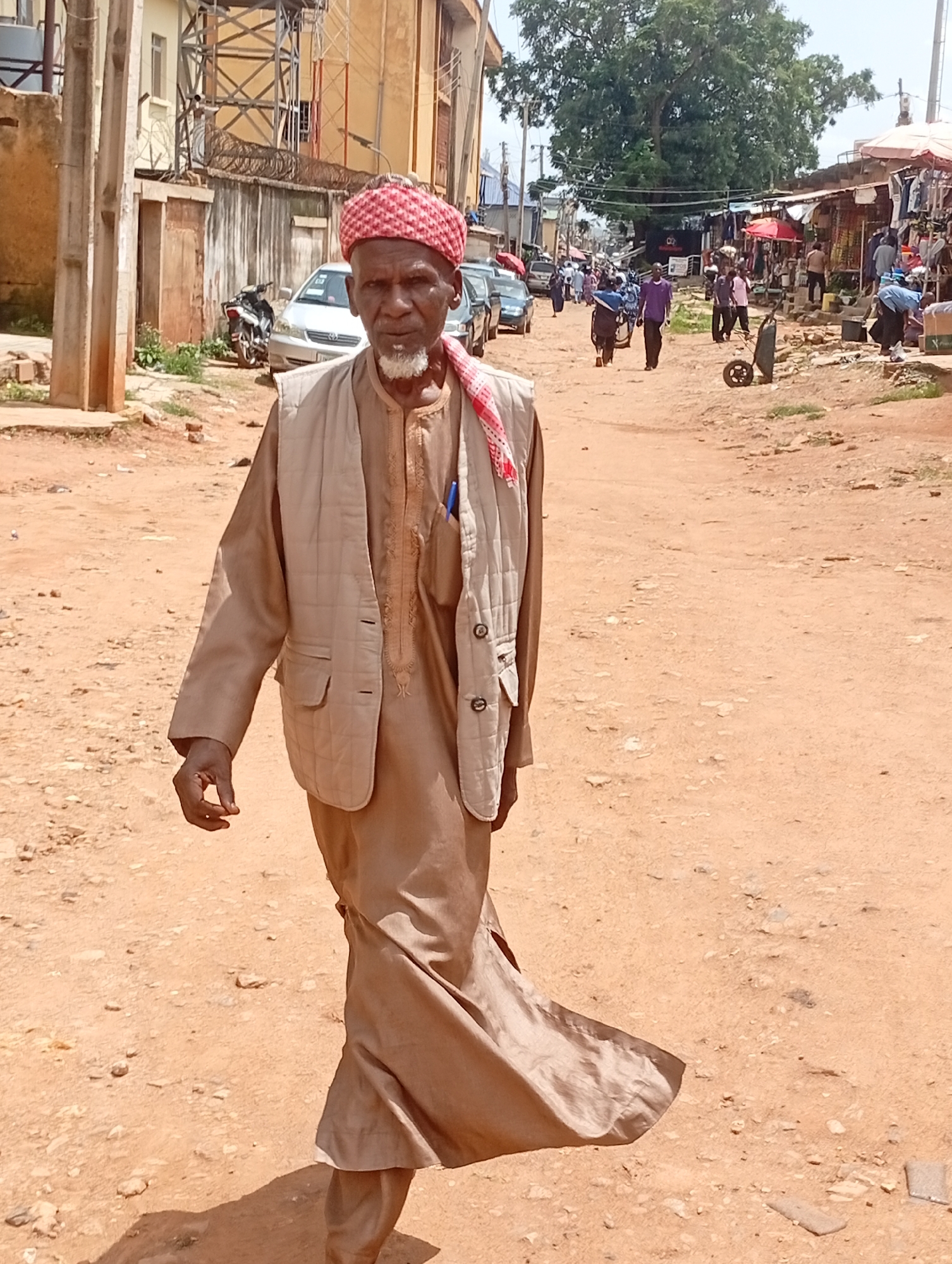 An elderly Black man wearing a traditional tan robe and a light beige vest, with a red and white patterned headscarf, walks on a dirt road in an outdoor marketplace. He has a white beard and appears to be looking forward. Behind him, other people are visible, some walking and others standing near market stalls. Cars are parked along the side of the road, and buildings line the street. The environment appears dry and dusty, consistent with the climate of Jos, Nigeria. The scene depicts everyday life in a bustling urban market.
