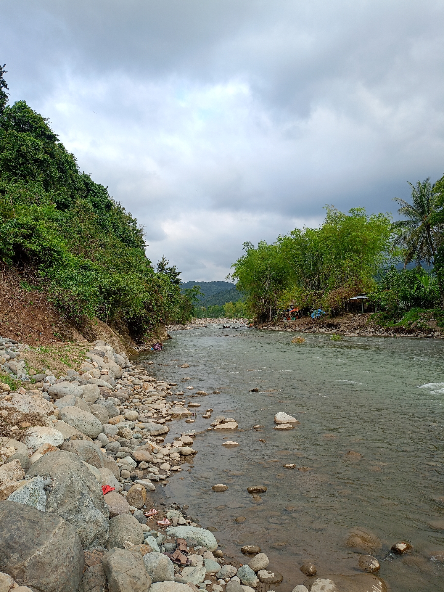 The image depicts a river in Meurah, Indonesia, under a cloudy sky. The river flows from the distance towards the viewer. The left bank is lined with large rocks and boulders, transitioning to a dirt bank covered with dense green vegetation. On the right bank, there is more green vegetation, including bamboo trees and a palm tree. Small, rudimentary structures are visible amidst the vegetation. People are present along the river, with two figures near the vegetated left bank and others near the huts to the right. The water appears clear and shallow, with rocks visible beneath its surface.