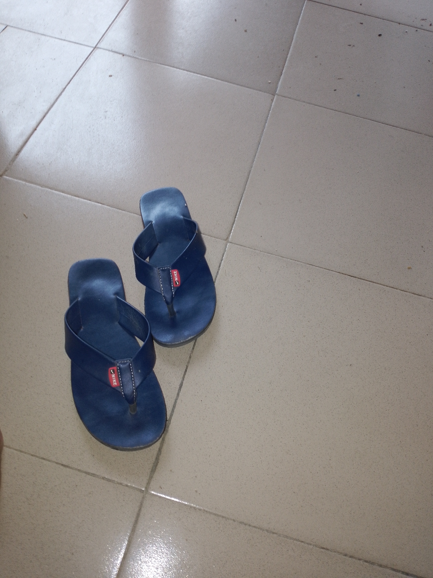A pair of dark blue Nike flip-flops are positioned on a beige tiled floor. The flip-flops are oriented with the soles facing downwards, and the straps are visible. The floor appears to be indoors, possibly in a home or public building, with visible grout lines between the tiles. The lighting suggests it is daytime, and there are some minor specks of debris scattered on the floor.