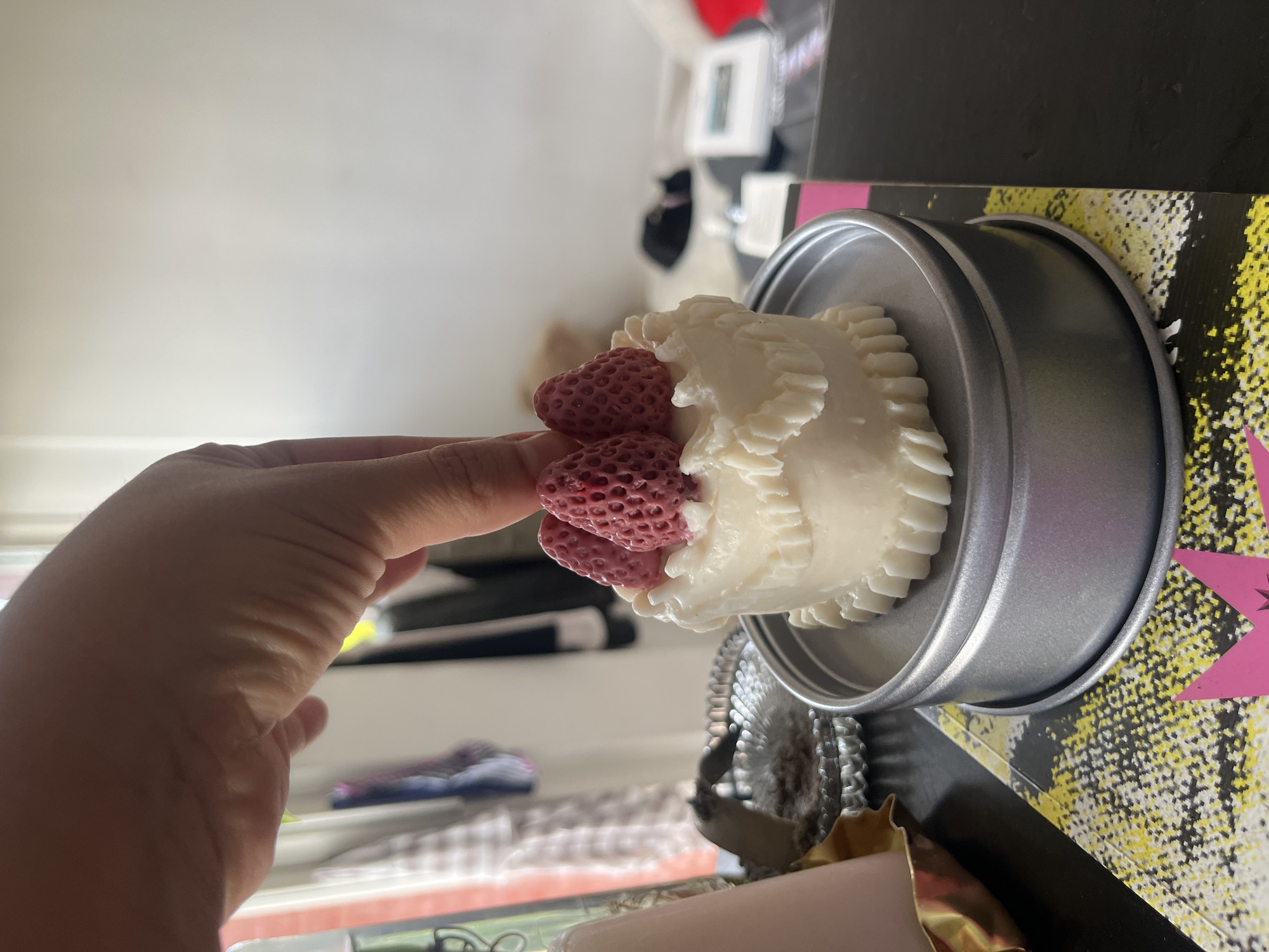 A human hand, light-skinned, is positioned centrally over a white, multi-layered, cake-shaped candle, appearing to adjust or place three deep pink, textured, strawberry-shaped objects on its top. The candle rests within a round, silver-colored metal tin. The tin is situated on a surface covered by a flat item, possibly a book or mat, displaying a yellow, black, and white speckled pattern with a fuchsia, pointed crown-like design visible at the lower left. To the immediate left, a peach-colored cylindrical object is partially visible within a golden, wavy-edged holder, adjacent to a round, reflective, textured metal tray. The background, largely out of focus, reveals an indoor setting with a brightly lit, plain light-colored wall, obscured hanging garments, and indistinct objects including a dark shape on a light surface and various rectangular items. The scene takes place within an interior room in Buenos Aires, Argentina.