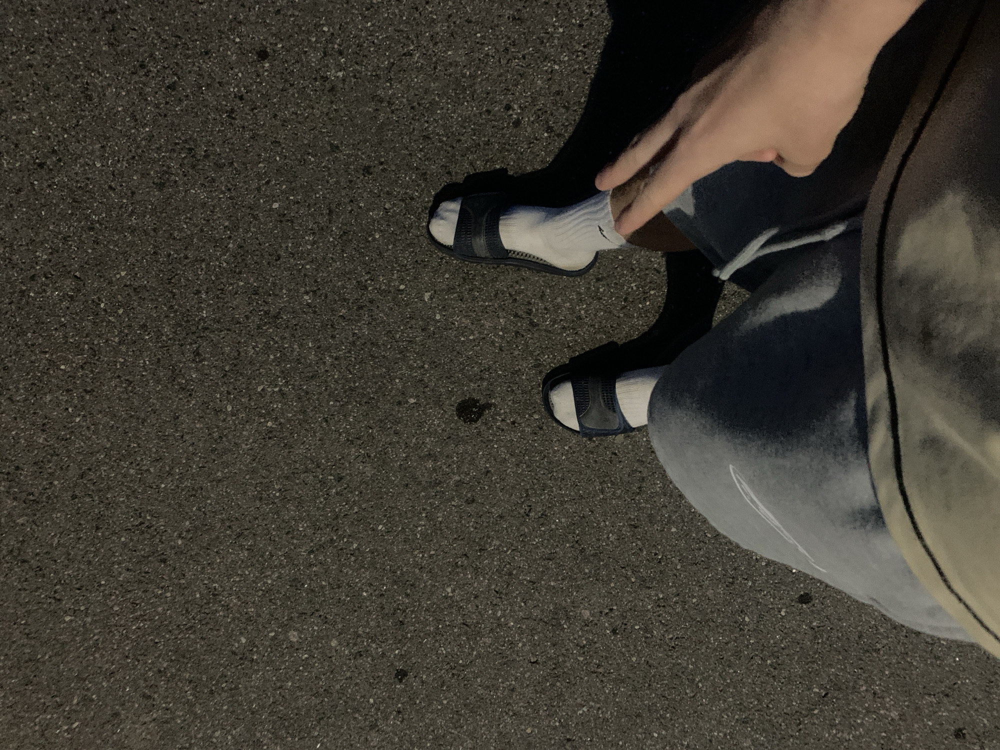 A first-person perspective shot shows a person standing on asphalt at night. The asphalt is dark gray and textured, with small white and gray stones visible. There are a few dark spots on the asphalt, possibly from a spill. The person is wearing light blue sweatpants, white socks, and dark blue slide sandals. One foot is clearly visible, and the other is partially visible. The person's right hand is in the foreground, extended towards the camera with two fingers slightly raised, forming a peace sign. The lighting suggests it is nighttime, with shadows cast by the person and their feet. The scene is devoid of any specific landmarks or identifying features that would indicate a precise location within the city of Minsk, Belarus.