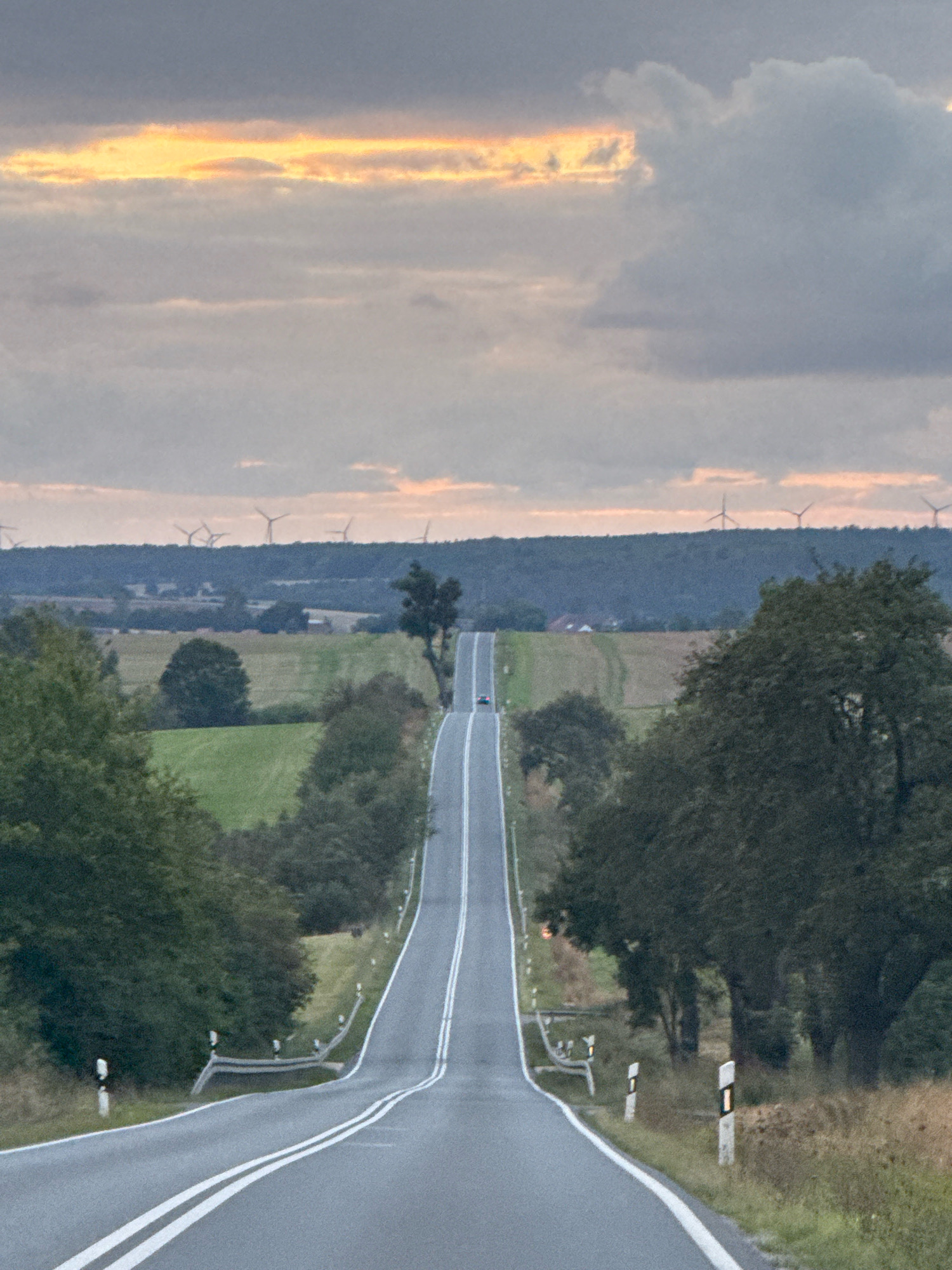 The image depicts a scenic rural landscape on a cloudy day, likely in the vicinity of Greußen, Germany, given the context. A paved road with double white lines curves into the distance, ascending a gentle hill. On either side of the road are grassy verges and mature trees, with fields and rolling hills visible in the background. In the far distance, a line of wind turbines dots the horizon, set against a muted sky with streaks of orange and yellow light breaking through the clouds. A single dark-colored car is visible on the road, heading uphill. The overall atmosphere is tranquil and pastoral.