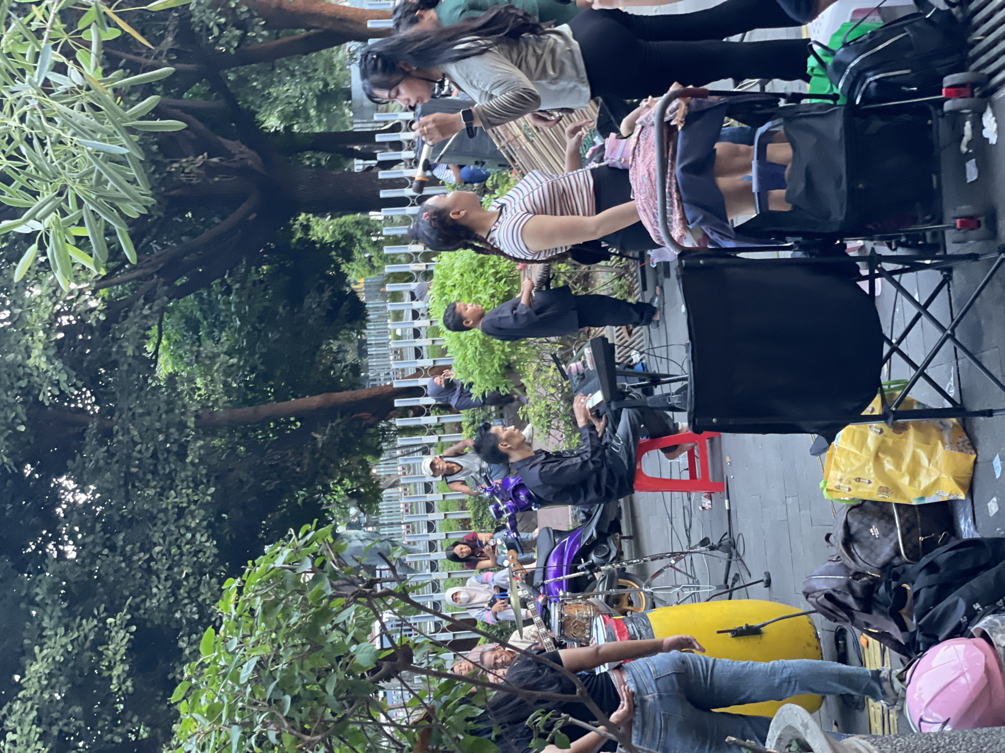 A group of people are gathered outdoors, likely in a park or public square, with a band performing. The scene is set in the city of West Jakarta, Indonesia.
In the foreground, a woman in blue jeans and a black top is standing, facing away from the camera. To her left, a large yellow drum is visible. Behind her, a keyboard player is seated on a red stool, playing a keyboard. To the right of the keyboard player, a woman in a striped shirt is standing and looking towards the right, seemingly engaged in conversation. Further to the right, another woman, dressed in a light-colored top and black pants, is holding a microphone and singing. A stroller is positioned in front of her.
In the background, several people are seated on a low wall or ledge, observing the performance. Among them are individuals wearing headscarves and casual clothing. A purple motorcycle is parked behind the musicians. The environment is lush with greenery, with trees and bushes providing a natural backdrop. A metal fence is visible behind the seated spectators. The ground is paved with tiles. The overall atmosphere appears to be casual and festive, suggesting a public event or street performance.