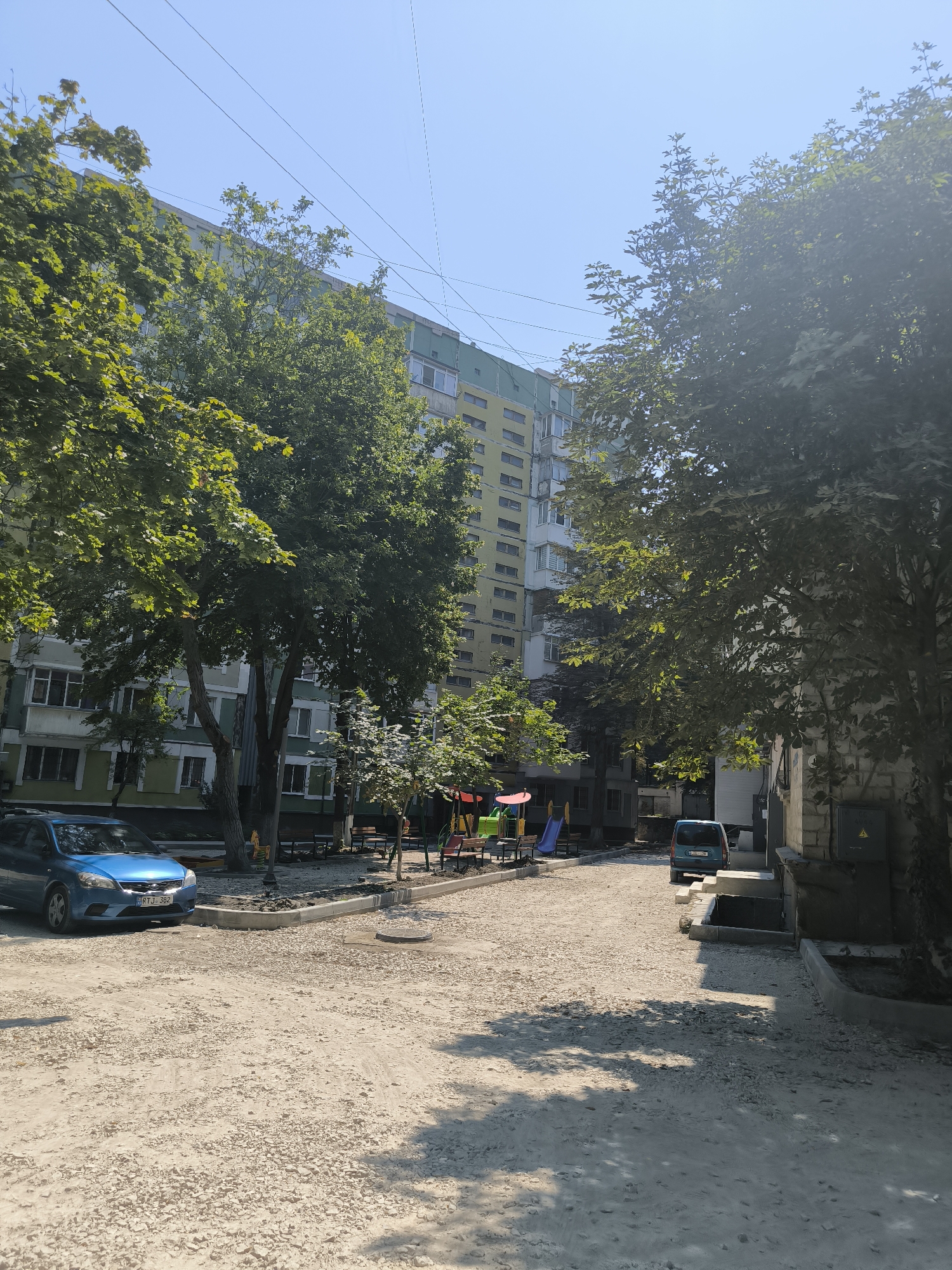 The image depicts a residential courtyard in Chișinău, Moldova, on a clear, sunny day. A blue car is parked in the foreground on a gravel-covered surface. Beyond the car, a children's playground with colorful slides and swings is visible. Several large trees provide shade, their green leaves contrasting with the light blue sky. In the background, a multi-story apartment building with a light green and yellow facade dominates the scene. Power lines are strung across the sky. A second, smaller blue vehicle is parked further into the courtyard, near the apartment building. The ground is a mixture of gravel and dirt, with some areas appearing to be under construction or recently disturbed. A manhole cover is visible in the foreground gravel.