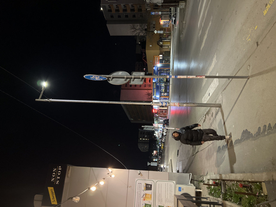 The scene depicts an urban street at night in Sofia, Bulgaria, specifically identifiable as the vicinity of Tsarigradsko Shose Blvd and Dobrich Street, near the Mladost 1 metro station area. A single male subject walks on the concrete sidewalk in the foreground, moving from left to right, with his gaze directed slightly towards the camera. He is dressed in a dark, hooded puffer jacket featuring a visible Nike logo on the left chest, dark patterned trousers, and light-colored athletic shoes. To the left, a white commercial building displays a yellow "Schweppes" sign and a "NON STOP" sign, with a vertical string of five illuminated spherical lights. Below these, a sign providing recycling information in Bulgarian is affixed above an outdoor air conditioning unit. A prominent grey metal streetlight pole stands in the mid-ground, fitted with a modern LED lamp that emits bright white light. Attached to this pole are multiple road signs, including a blue circular sign with a white bicycle symbol indicating a mandatory bicycle route. Across the asphalt road, a pedestrian crosswalk is marked by white stripes. Commercial buildings line the opposite side of the street, featuring illuminated storefronts for businesses such as an "Inbet" betting shop and an "Аптека" (pharmacy), with multi-story residential structures visible further in the background. Overhead utility wires traverse the dark, clear night sky. Several vehicles, including parked cars and cars in motion, are discernible along the street. The ground appears dry, and artificial light sources illuminate the scene, casting shadows from the pedestrian and the streetlight pole.