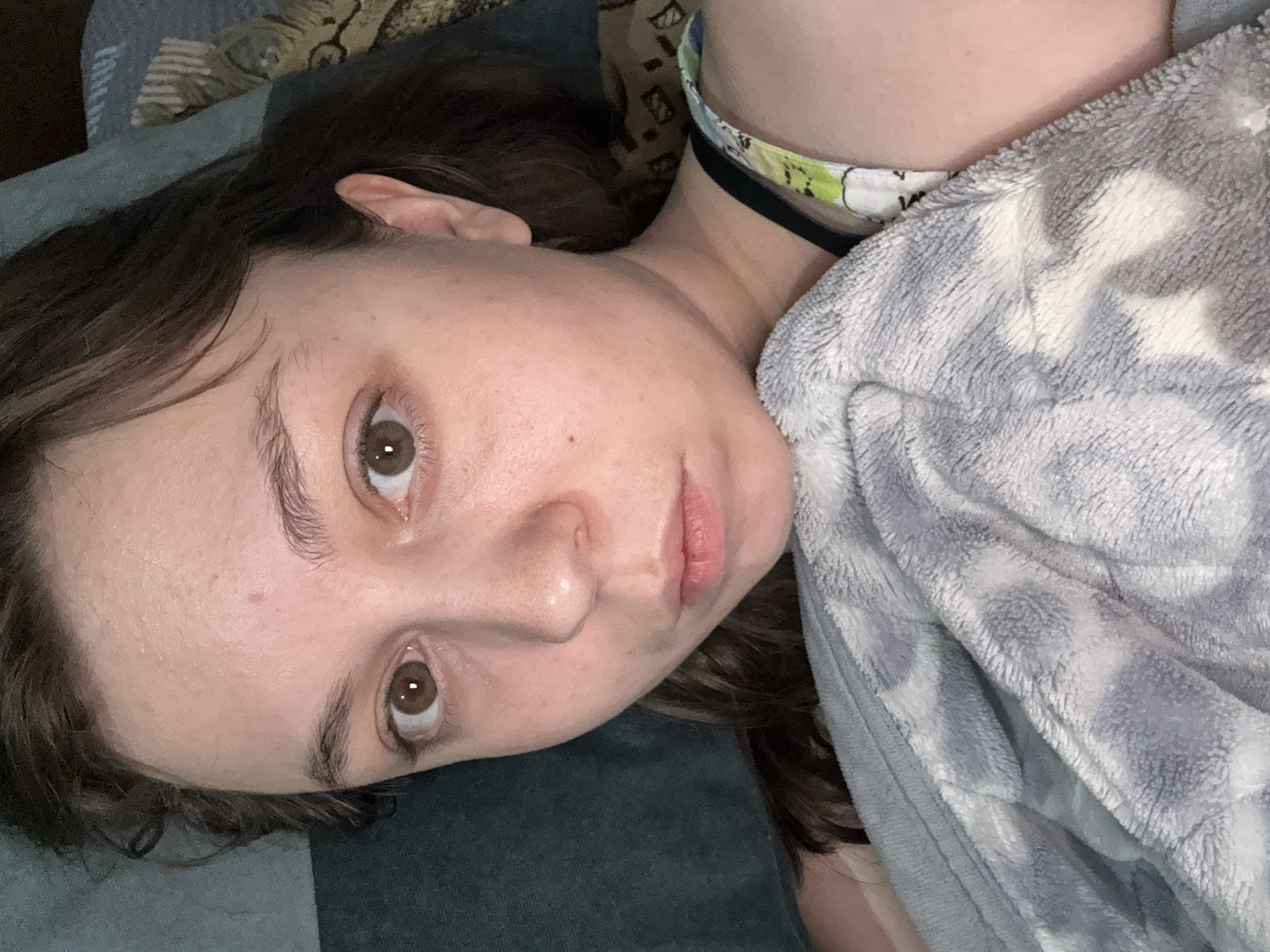 The image shows a close-up, over-the-shoulder selfie of a young woman lying down, possibly in bed. She has brown hair, brown eyes, and fair skin. She is wearing a patterned strap, possibly from a tank top or bra, and is covered with a soft, grey and white patterned blanket. The background appears to be a dimly lit room with textures suggesting bedding and possibly a wall. There is no discernible information in the image that would allow for identification of a specific location within Shymkent, Kazakhstan.