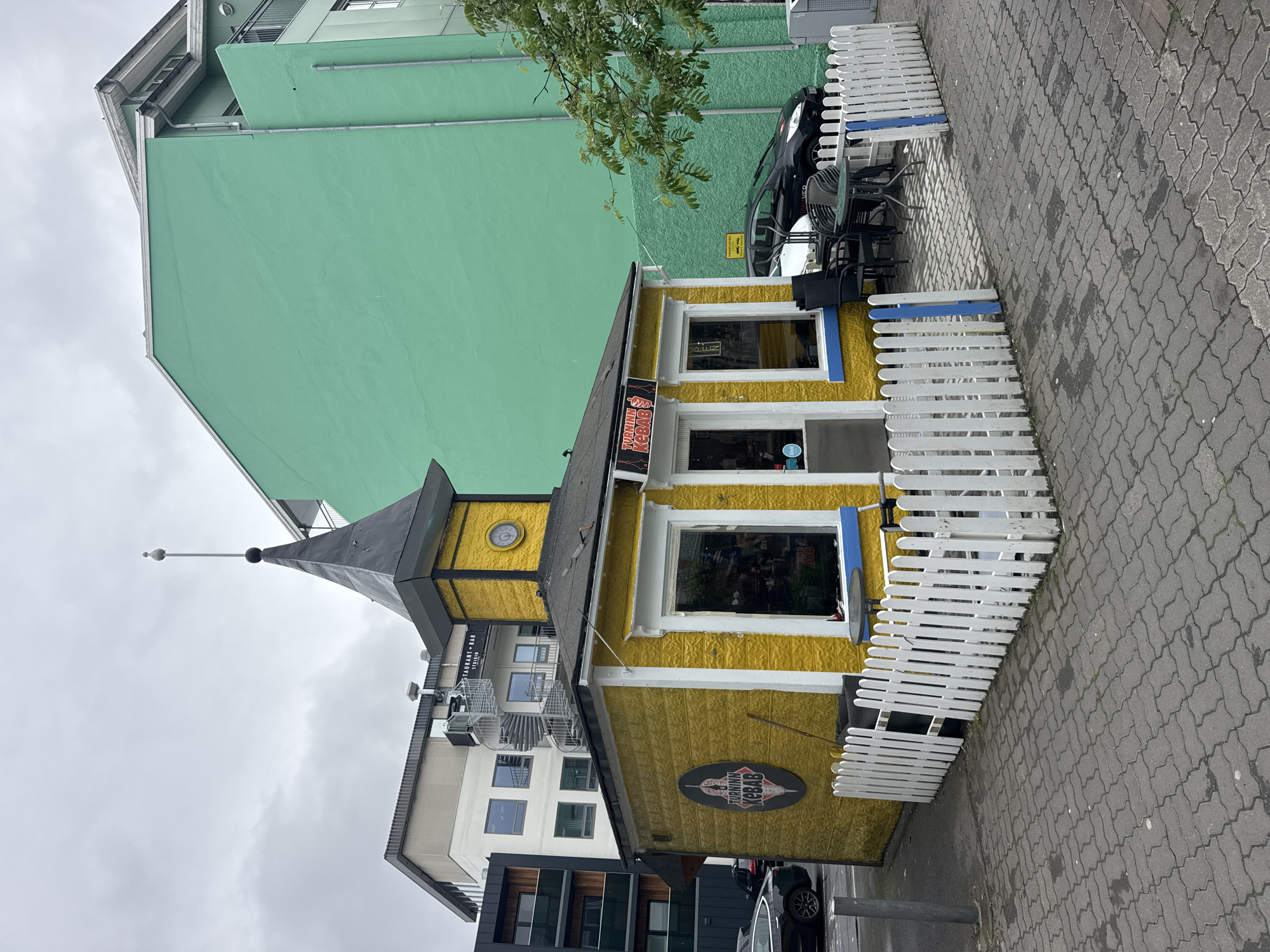 The image captures an urban street scene, focusing on a vibrant yellow building with a distinctive dark grey pointed roof, almost spire-like, crowned with a finial. This small structure features white window frames with blue accents beneath the windows and a small round clock near the spire. A white picket fence extends along the front of the building, enclosing an outdoor seating area furnished with dark tables and chairs.
Adjacent to the yellow building and receding into the background is a larger, multi-story building, painted in a striking bright green, with parts of its facade in white and grey. A spiral staircase is visible on one side of this taller building. The foreground features a paved sidewalk made of interlocking grey pavers. A black car is partially visible, parked behind the white fence, and a tree branch with green leaves overhangs the scene.
The sky is overcast, indicating a cloudy daytime. No people are present. Several signs are visible on the yellow building: "TURNNIN KEBAB" appears on a sign above the door and on a circular sign featuring a kebab illustration on the lower left side. On the larger background building, partial text "RESTAURANT - BAR" and "STEINB" can be seen. The setting suggests a commercial area, consistent with the location context of Akureyri, Iceland.
