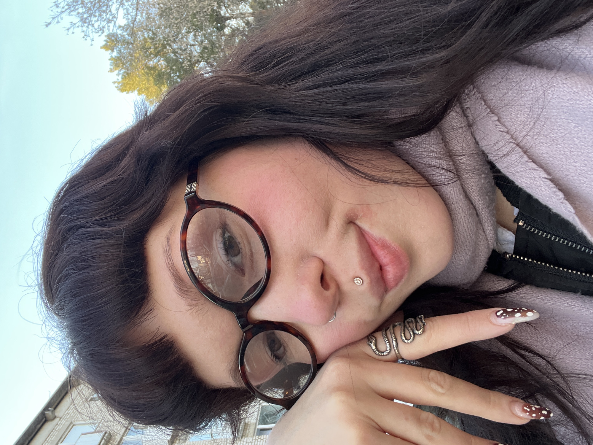 A selfie-style image shows a young woman with dark brown hair and fair skin lying outdoors. She is wearing eyeglasses with tortoise-shell frames and a lavender-colored hoodie. She has a lip piercing and a nose ring. Her nails are adorned with a brown and white spotted design, and she wears a snake ring on her finger. Her face is tilted, and she is looking towards the camera.
The background includes glimpses of a building and some trees, suggesting an outdoor location. The architecture, combined with the style of foliage, could potentially place the image in or near the city of Chaikovsky, Russia.