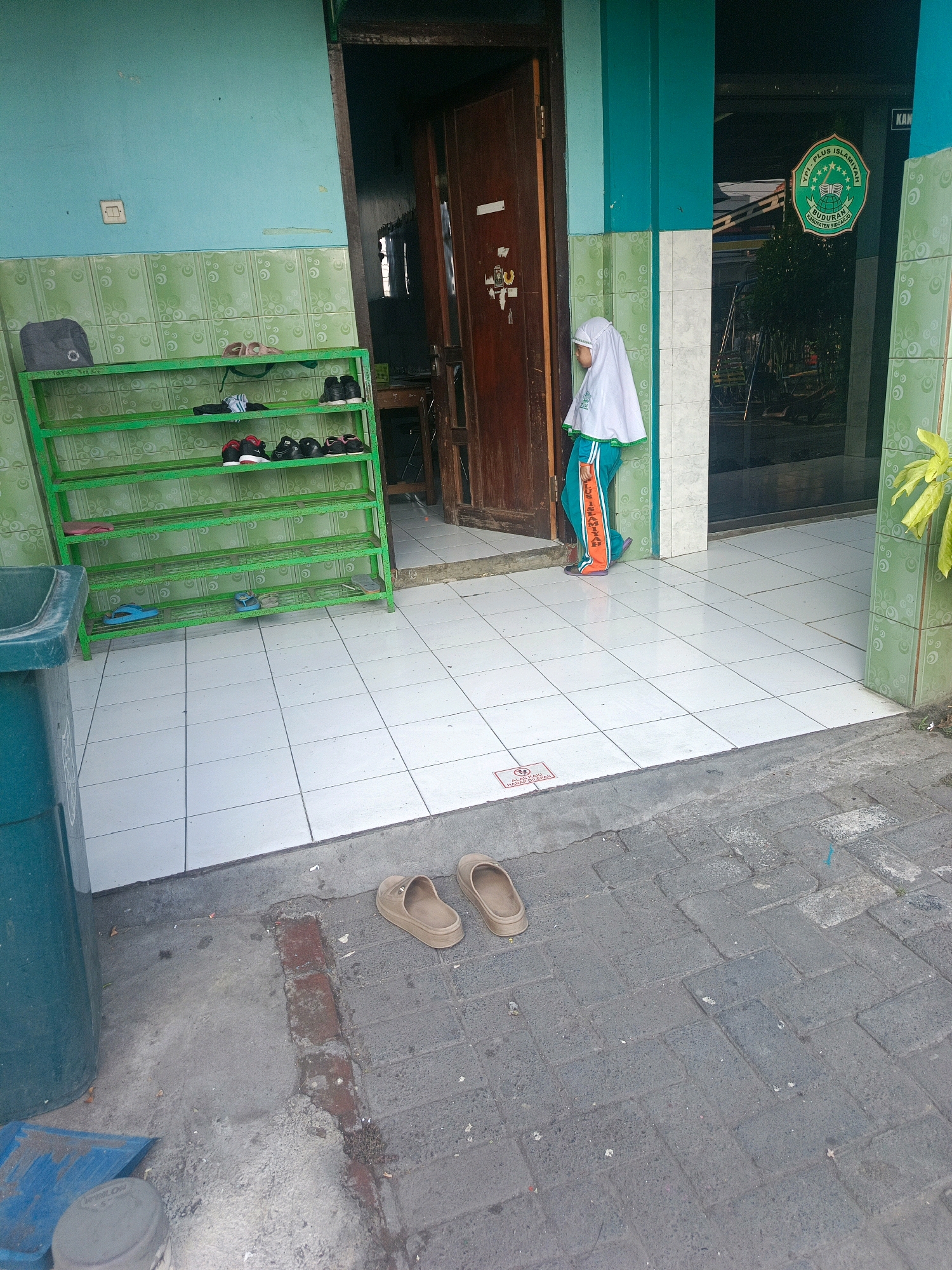 The image depicts an outdoor scene in front of a building, likely a school, in Banjarsari, Indonesia. In the foreground, two tan-colored sandals are placed on the paved ground. To the left, a large teal-colored trash bin and a blue dustpan with a grey lid are visible. A green shoe rack, filled with various pairs of shoes and sandals, stands against a light teal wall adorned with green patterned tiles. A child wearing a white hijab and an orange and green uniform is standing in a doorway to the right, looking towards the interior of the building. The doorway is framed by a wooden door and light teal and white tiled walls. Above the doorway, a circular green and white emblem with text is affixed to the wall. The ground is a mix of white tiled pavement and darker brick paving stones. The overall environment suggests a functional outdoor space associated with an educational institution.
