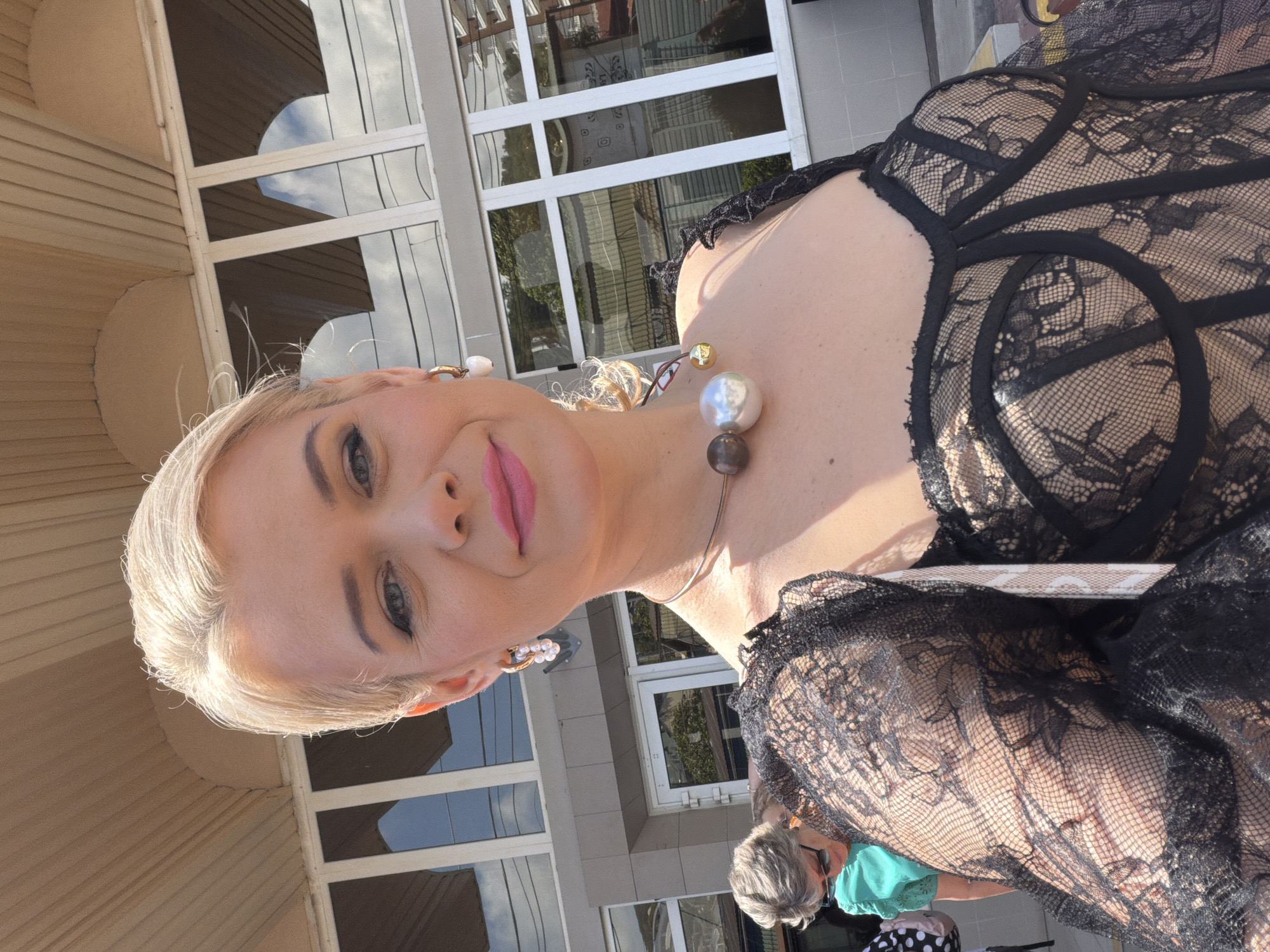 A woman is taking a selfie outdoors, possibly in Sochi, Russia. She has blonde hair and is wearing a black lace top. A necklace with large, spherical beads adorns her neck. In the background, a building with large windows and striped siding is visible, suggesting an urban or suburban setting. The sunlight indicates it is daytime, and the clear sky hints at good weather. The woman's pose and direct gaze at the camera suggest she is aware of being photographed.