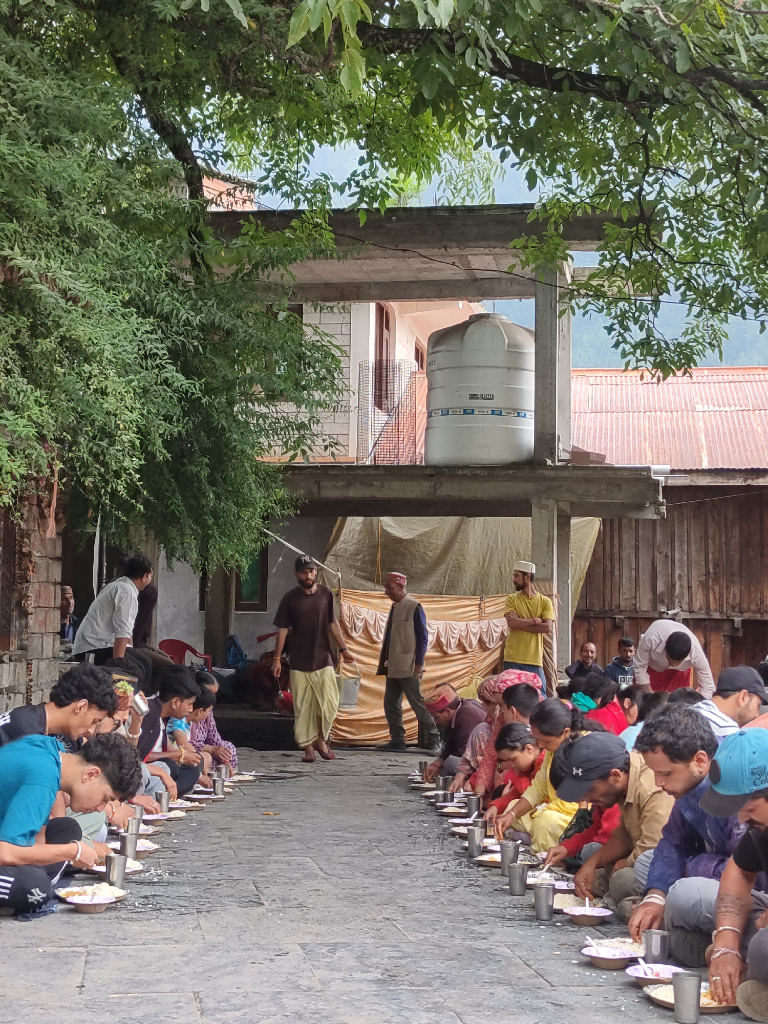 In an outdoor courtyard setting, a group of people are seated on the ground, forming two long lines, facing each other. They are engaged in a communal meal, with plates of food and metal cups laid out before each individual. The people are dressed in a mix of traditional and contemporary clothing.
In the background, a two-story structure is visible, with a large white water tank on the upper level. The building appears to be made of brick and concrete. Adjacent to this structure is a wooden building with a corrugated metal roof. A large tarp is draped behind the diners, serving as a backdrop.
Several trees with dense green foliage overhang the scene, providing shade. The ground is paved with stone. The overall atmosphere suggests a gathering or a religious or cultural event. The location is described as "Unknown, Unknown," implying it is not a well-known or specific landmark.