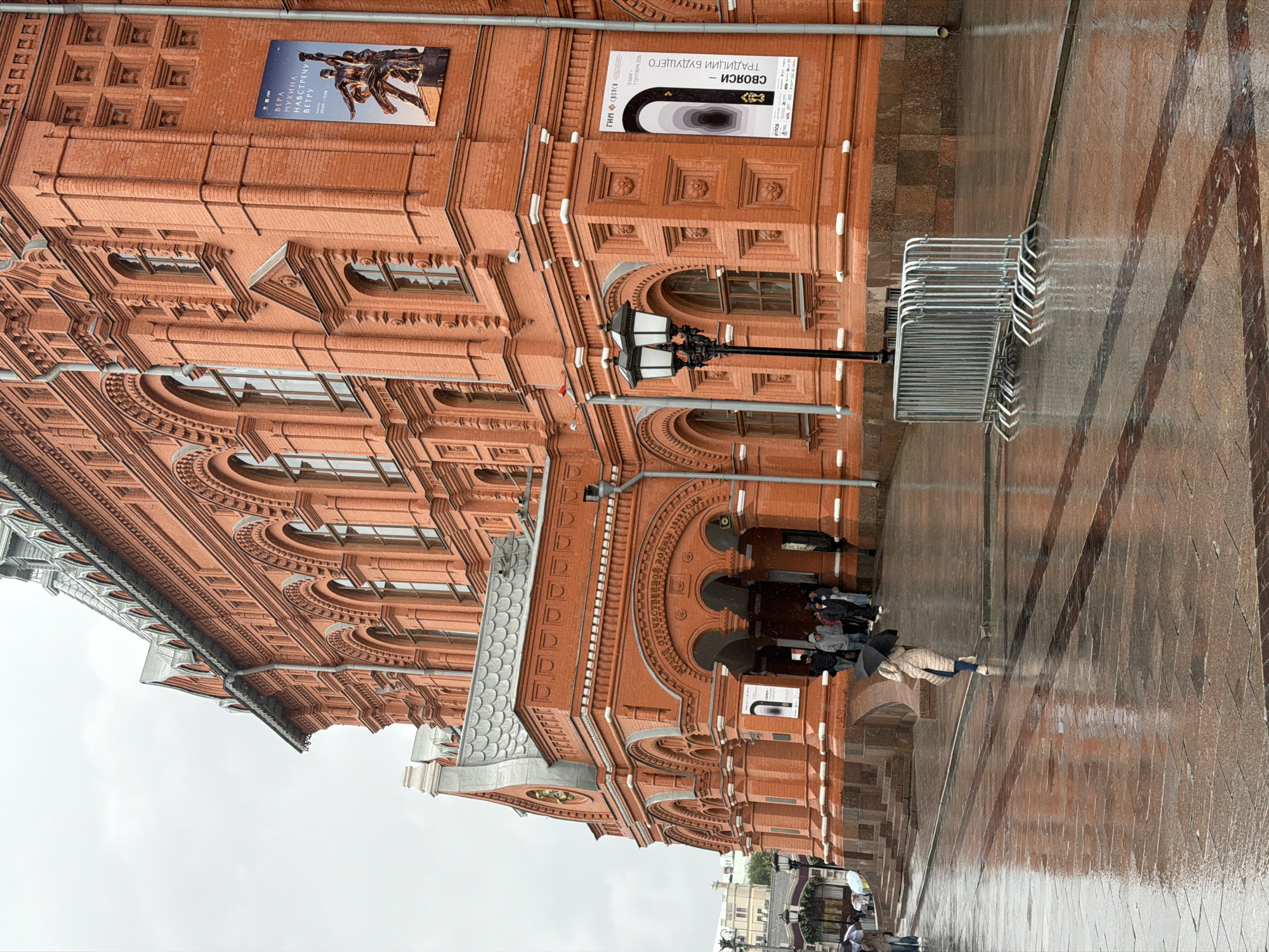 The image depicts the exterior of the State Historical Museum in Red Square, Moscow, Russia. It is a rainy day, and the pavement is wet and reflective. Several people are visible, some with umbrellas, walking near the entrance of the museum. The architecture of the museum is characterized by its distinct red brick facade and arched windows. Two large posters are affixed to the exterior wall of the building. In the foreground, a metal barrier is set up on the wet pavement. The sky is overcast, contributing to the subdued lighting of the scene.