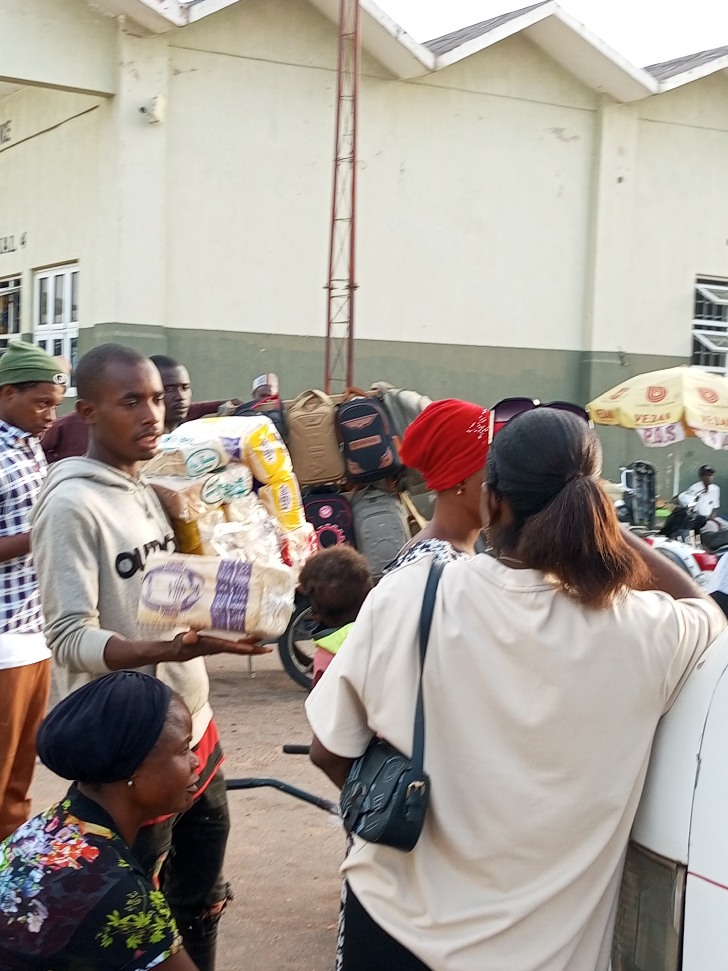 In Gombe, Nigeria, a group of people are gathered outdoors, near a building with a light-colored facade and a sloping roof. The central figure, a young man wearing a grey hoodie, is holding a stack of bread loaves. Several other individuals are visible, including a woman in the foreground with a black headscarf and a floral-patterned top, and two women behind the bread-holder, one wearing a red headscarf. Backpacks and a yellow umbrella are present in the background, suggesting a travel or market setting. The ground is paved with asphalt.