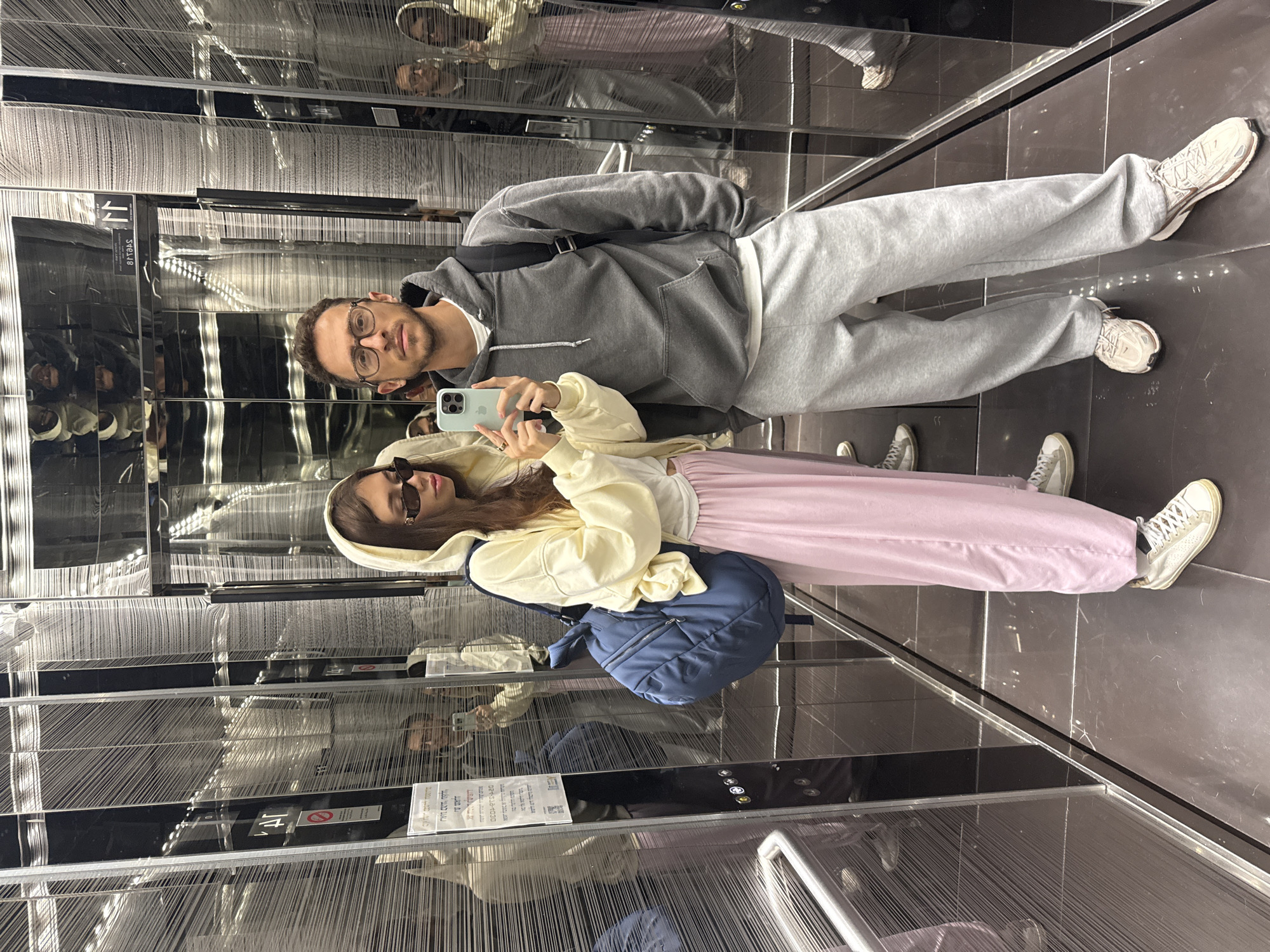 A man and a woman are taking a picture of themselves in an elevator with mirrored walls. The elevator appears to be located in a modern building in Tel-Aviv, Israel. The man is wearing glasses, a grey hoodie, and grey sweatpants. The woman is wearing sunglasses, a light yellow hoodie, and pink sweatpants. She is carrying a blue backpack. The elevator walls have a decorative design of vertical lines. The floor is made of dark tiles.