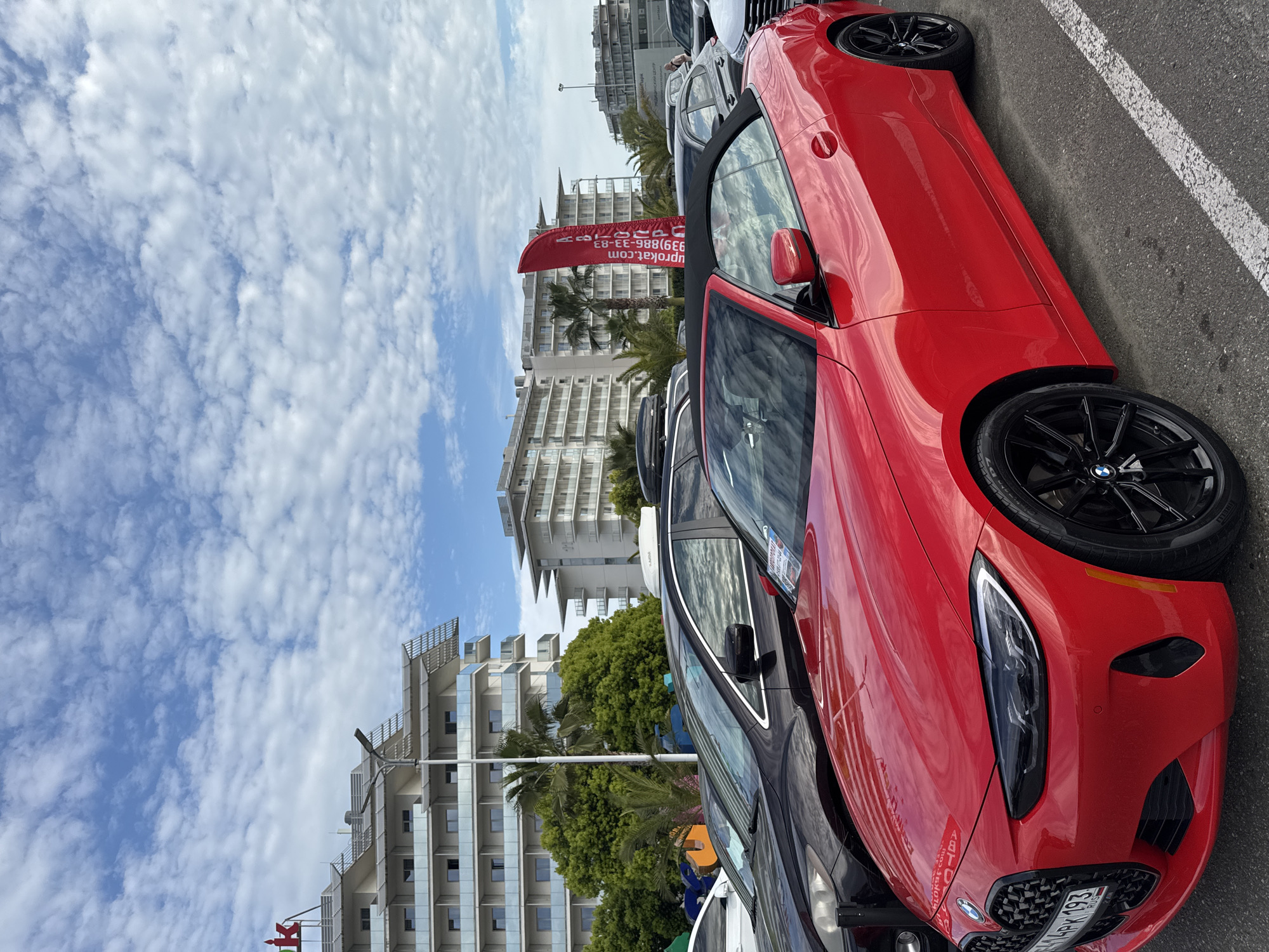 The image captures a street scene in Sochi, Russia, showcasing a row of parked cars against a backdrop of modern, multi-story buildings. A red BMW convertible dominates the foreground. Adjacent to the BMW is a dark-colored vehicle. A red flag with Cyrillic writing is visible near the buildings. The sky above is partly cloudy. The buildings exhibit contemporary architecture with glass and geometric features. Palm trees are scattered among the buildings, indicating a coastal environment. The scene appears to be an outdoor, daytime setting. The BMW is partially in focus, drawing the viewer's attention to the car's front end and wheel. The parking space is marked with white lines.